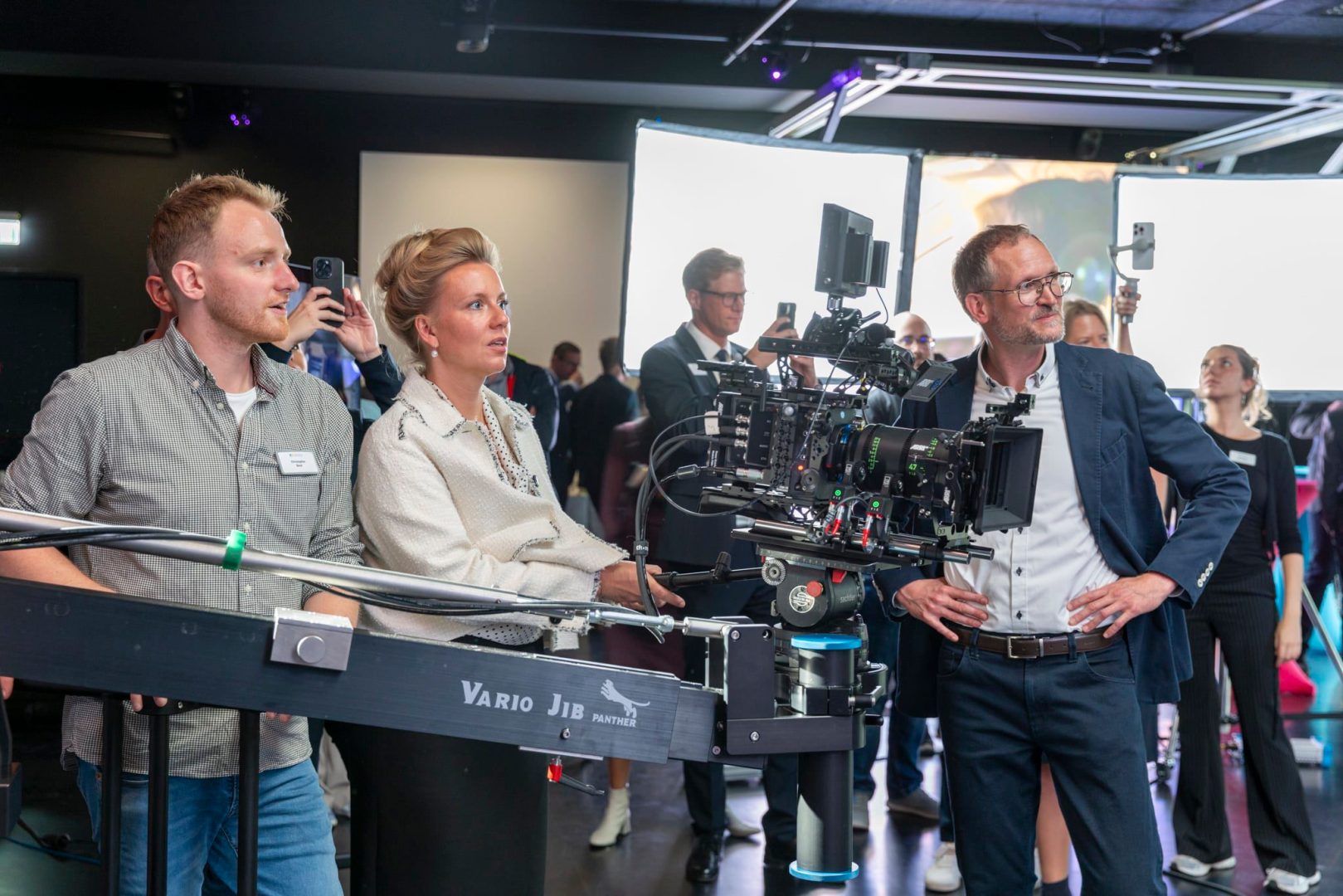 a group of people observing a film production setup with a camera rig in focus two individuals in the foreground one with a clipboard look attentively while others watch from the background in a modern studio environment digital production A group of people observing a film production setup, with a camera rig in focus. Two individuals in the foreground, one with a clipboard, look attentively, while others watch from the background in a modern studio environment.