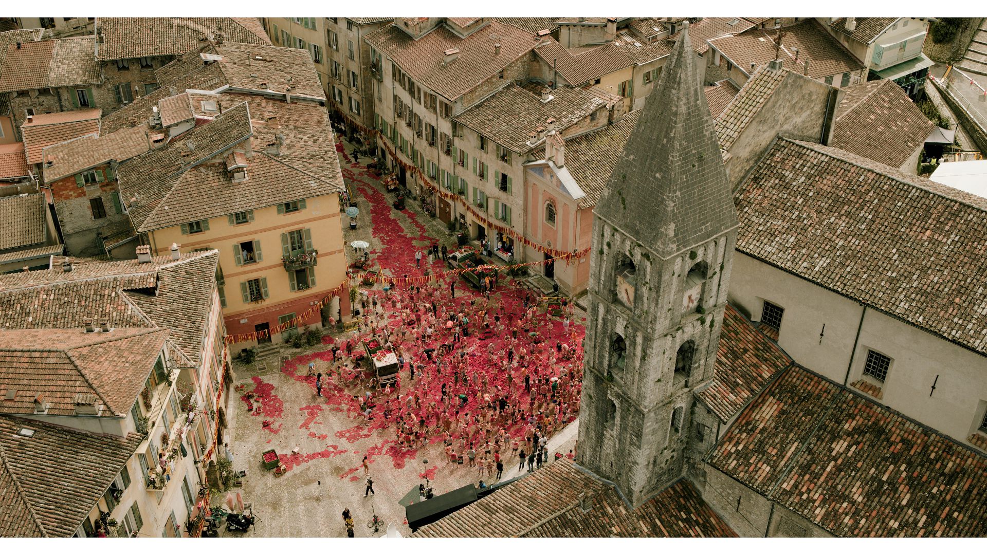 An aerial view of a lively plaza filled with people celebrating, surrounded by historic buildings and a church tower. The ground is covered in bright red fabric or paper, creating a vibrant scene.