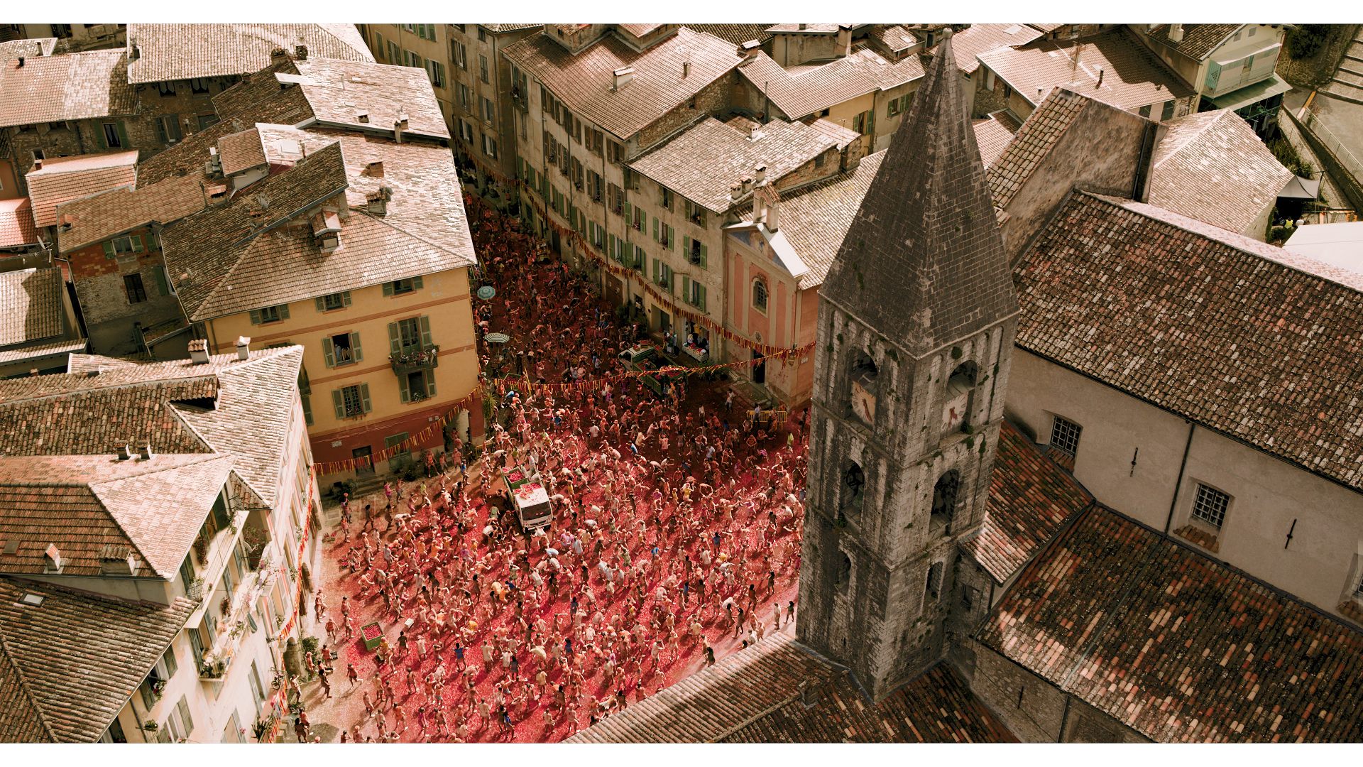 Aerial view of a large crowd wearing red in a narrow street surrounded by buildings and a church tower. The crowd appears to be celebrating an event.