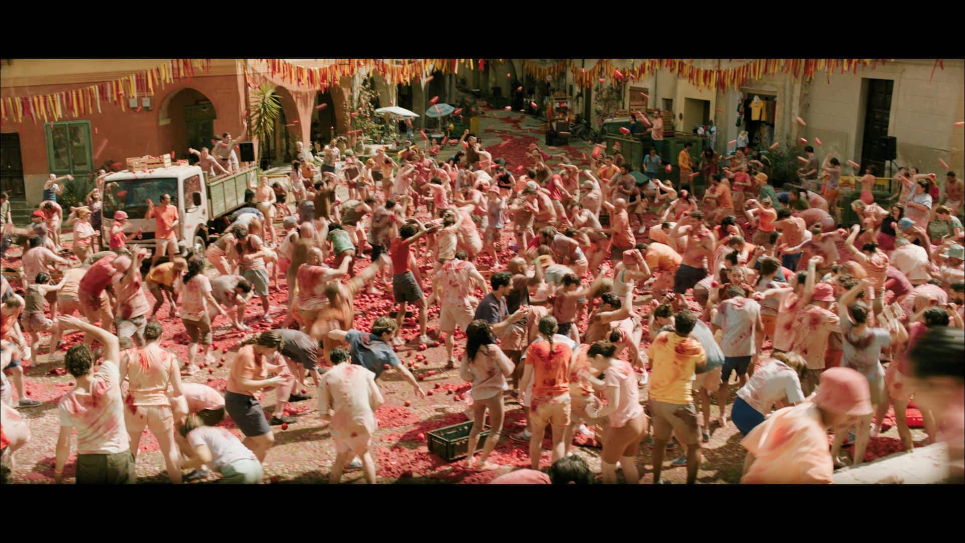 A large crowd of people participating in a festive event, surrounded by discarded tomatoes. Many participants are shirtless and covered in red pulp, with decorations hanging overhead in a lively atmosphere.