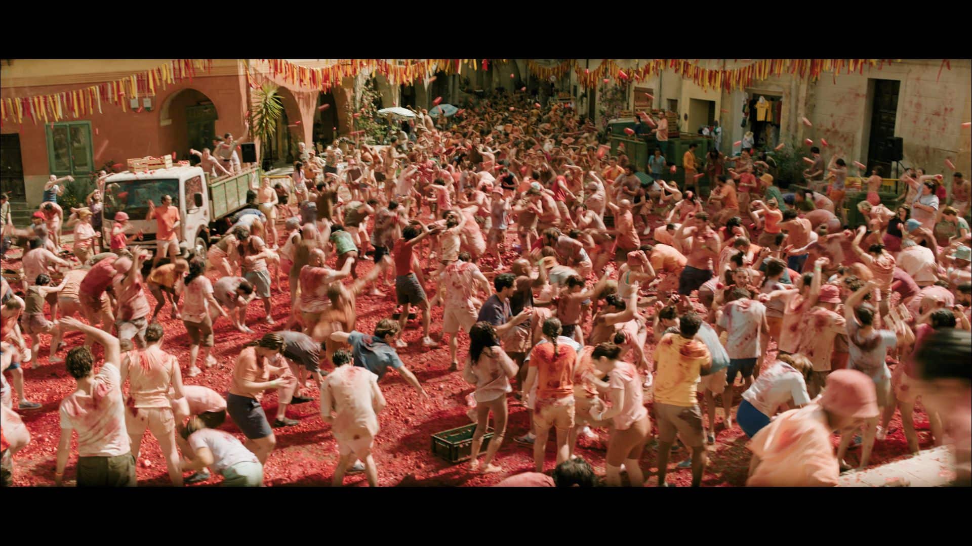 A large crowd of people covered in red pulp, participating in a festival in a street decorated with colorful flags. Many individuals are throwing tomatoes, creating a lively scene filled with activity.