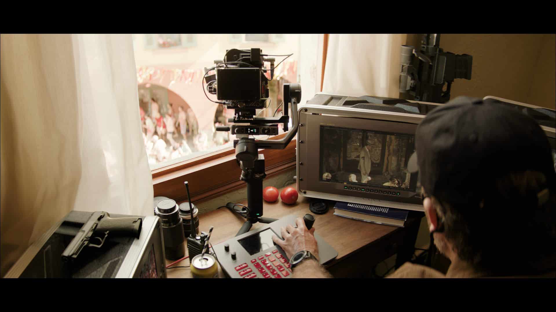 a person operating a camera and monitor setup inside a room two apples are on the desk along with a beverage can and tools a window shows an outside view with blurred figures and buildings digital production A person operating a camera and monitor setup inside a room. Two apples are on the desk along with a beverage can and tools. A window shows an outside view with blurred figures and buildings.