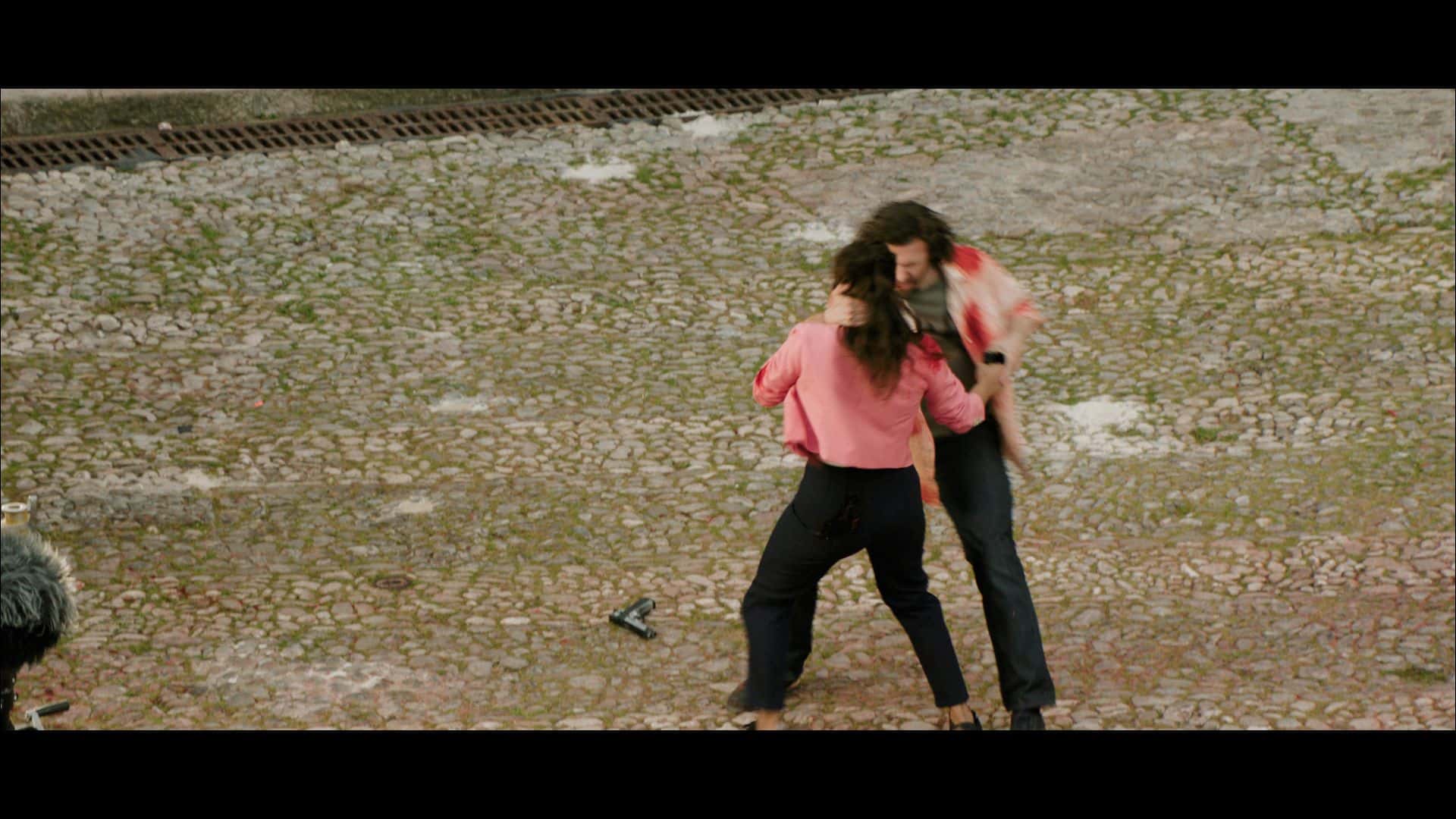 two people are engaged in a physical altercation on a cobblestone surface one person is wearing a pink top and black pants while the other is in a red and black shirt with dark pants a microphone can be seen in the foreground digital production Two people are engaged in a physical altercation on a cobblestone surface. One person is wearing a pink top and black pants, while the other is in a red and black shirt with dark pants. A microphone can be seen in the foreground.