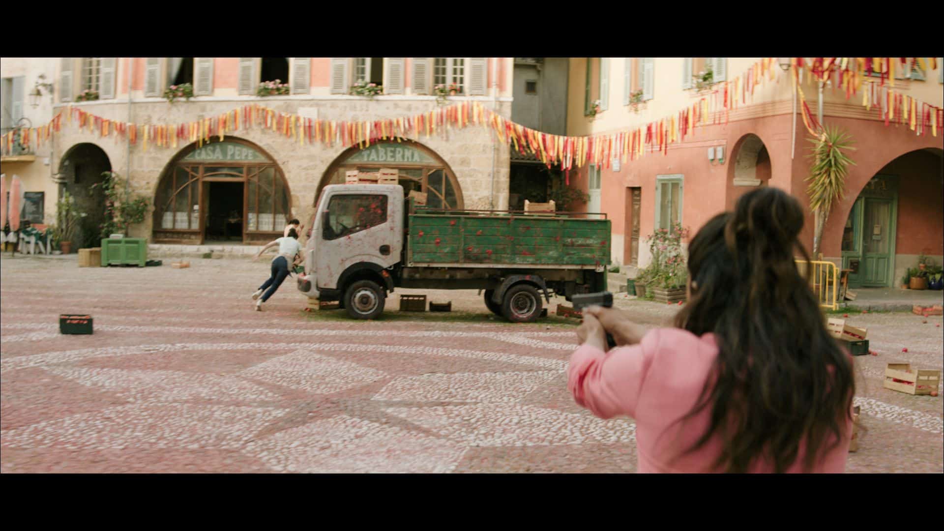 A woman with a gun is aiming at a man running away near a green truck in a decorated plaza. Colorful flags hang above the area, and there are crates on the ground.