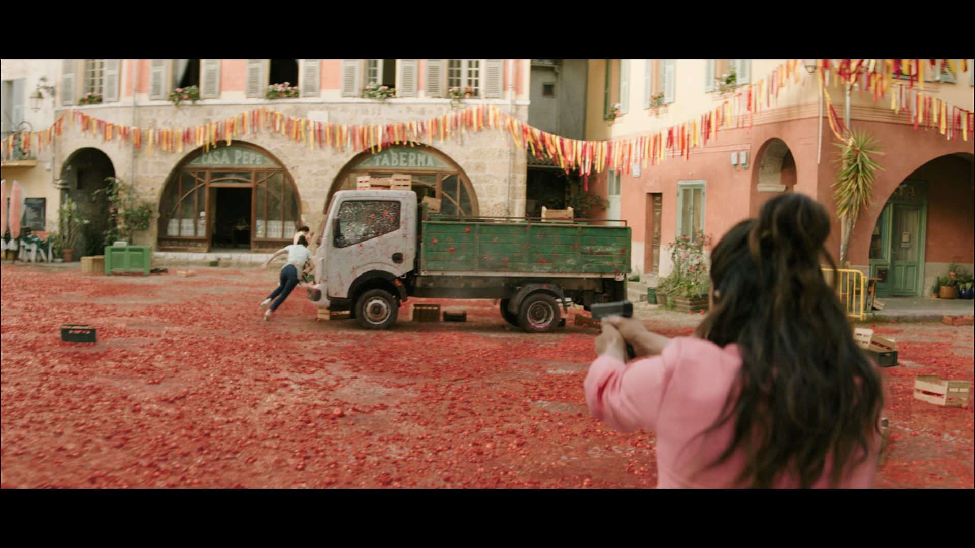 A person running on a street covered with tomatoes, while another person holds a gun. In the background, a green truck is parked near buildings decorated with colorful banners.