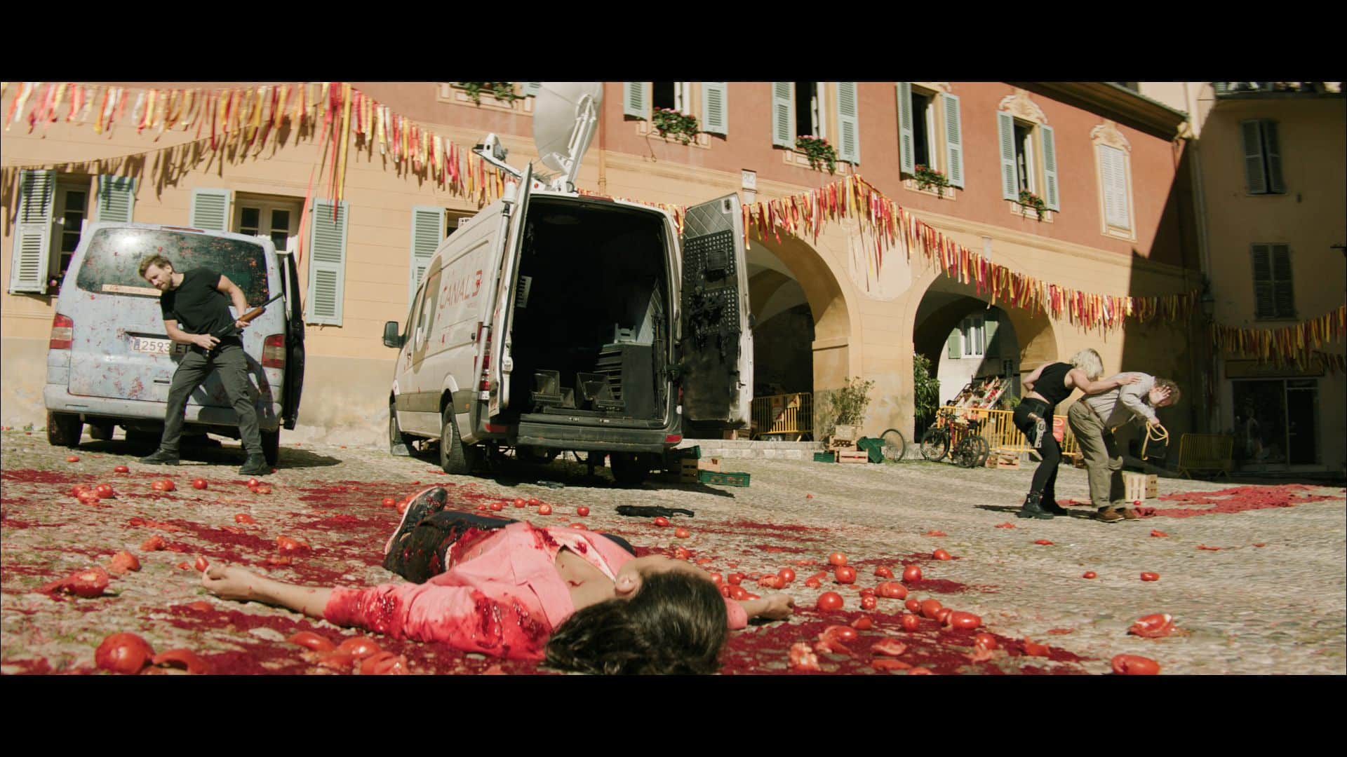 A street scene with two vehicles parked. A person lying on the ground in a red shirt. Another person is bending over nearby, while another stands with seeming urgency. The ground is scattered with red items, resembling tomatoes.