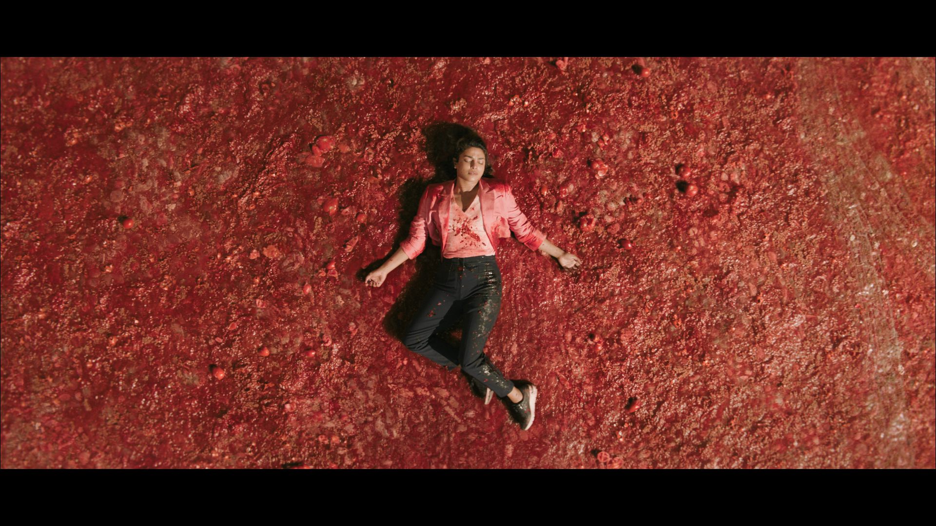 A person lying on a red textured surface, wearing a pink shirt and jeans, with arms outstretched. The background appears to be a field or surface with a red substance.