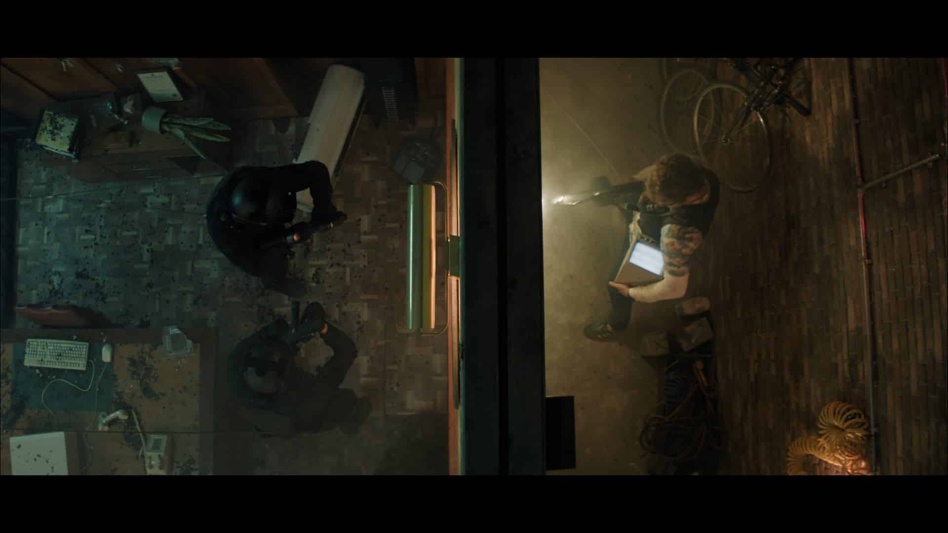 An overhead view of a room showing two groups of people. One group includes two individuals in black clothing holding guns, and the other group consists of two people, one with a light and the other holding a device, in a tense situation.