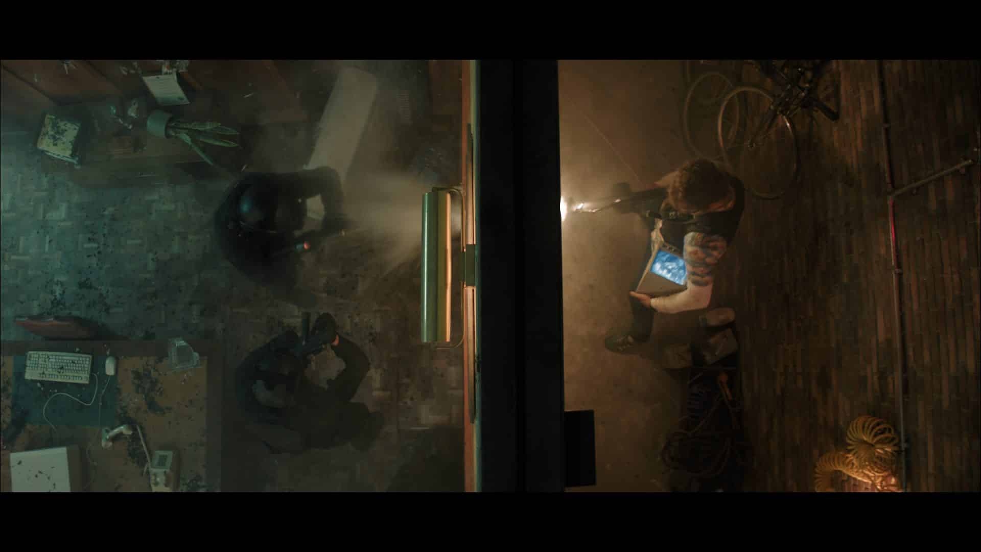 Aerial view of a dimly lit room with two people holding flashlights. One person is standing while the other is crouched on the floor. Tools and debris are scattered around the floor, with wires visible in the background.