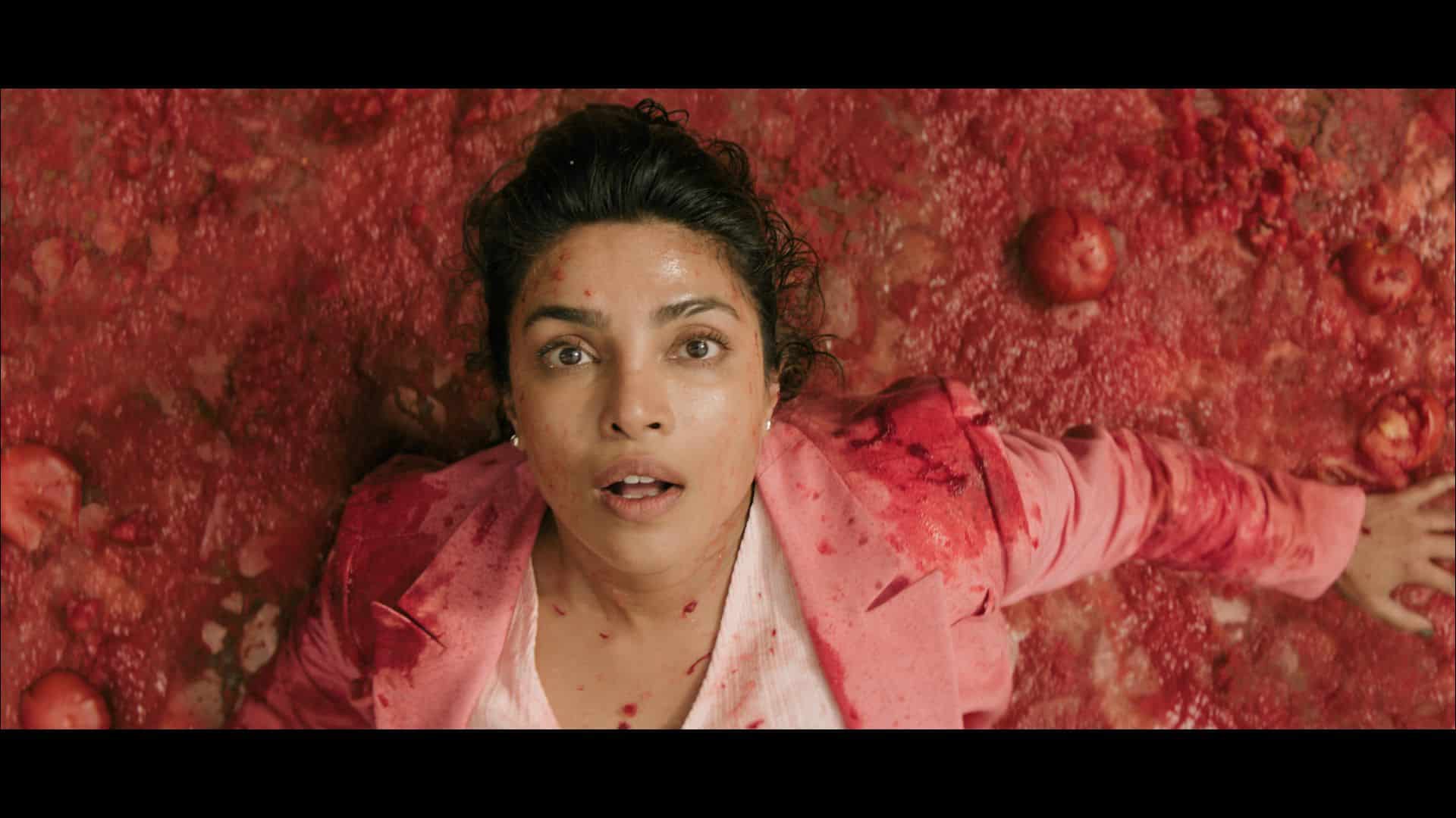 A woman lying on a surface covered with red substance, surrounded by scattered round objects. Her expression is neutral, and she is wearing a pink jacket over a white top.