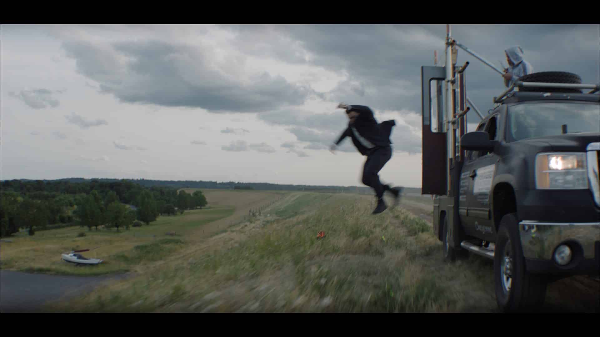 a person in a black outfit jumps from a dirt road next to a pickup truck the background features grassy terrain and a cloudy sky digital production A person in a black outfit jumps from a dirt road next to a pickup truck. The background features grassy terrain and a cloudy sky.