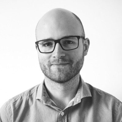 a man with a shaved head and glasses smiling at the camera he is wearing a collared shirt and has a light expression the background is plain white digital production A man with a shaved head and glasses smiling at the camera. He is wearing a collared shirt and has a light expression. The background is plain white.