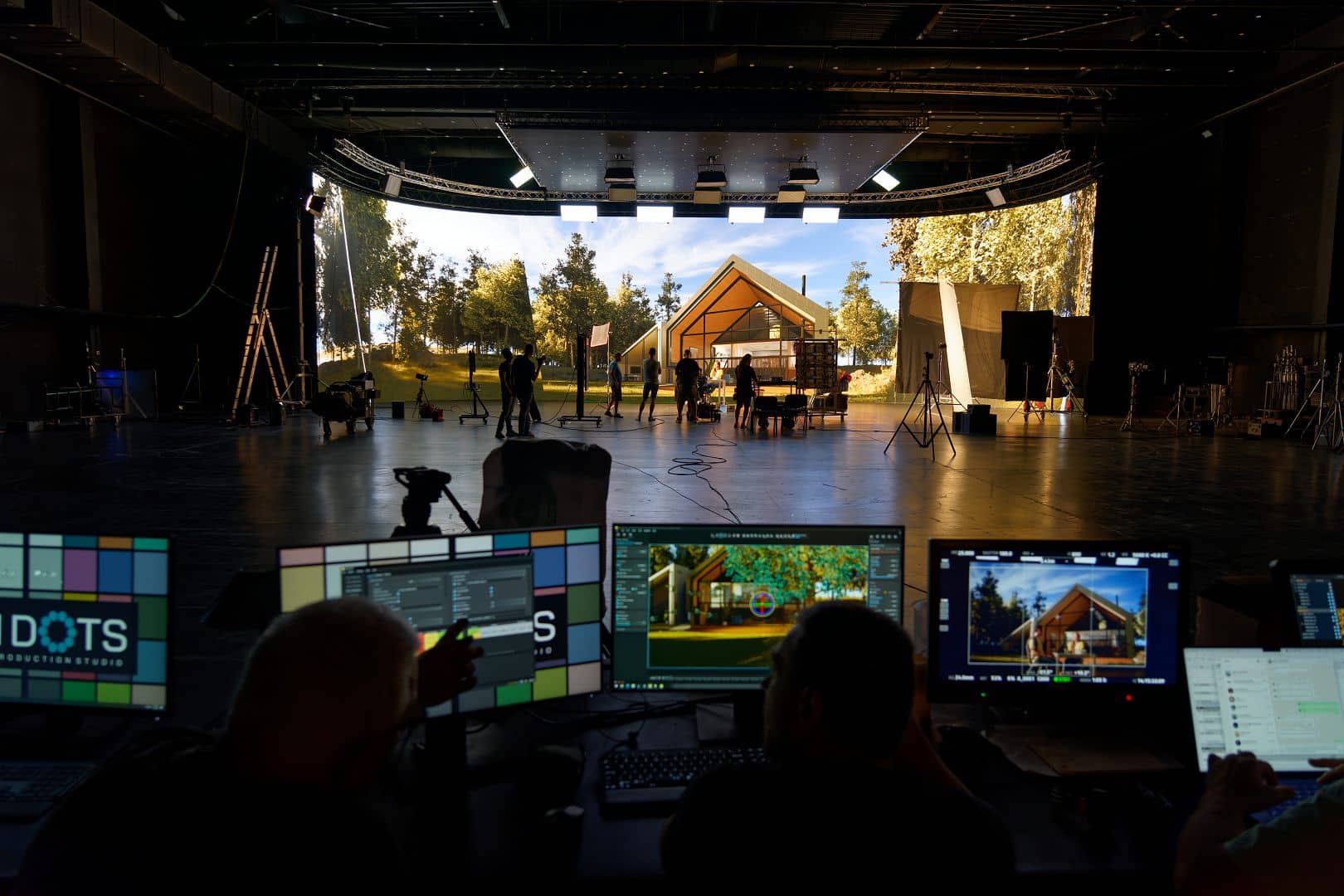 a film studio with two operators at computer monitors adjusting settings in the background a group of people are setting up equipment near a wooden structure surrounded by trees and outdoor lighting digital production A film studio with two operators at computer monitors, adjusting settings. In the background, a group of people are setting up equipment near a wooden structure surrounded by trees and outdoor lighting.