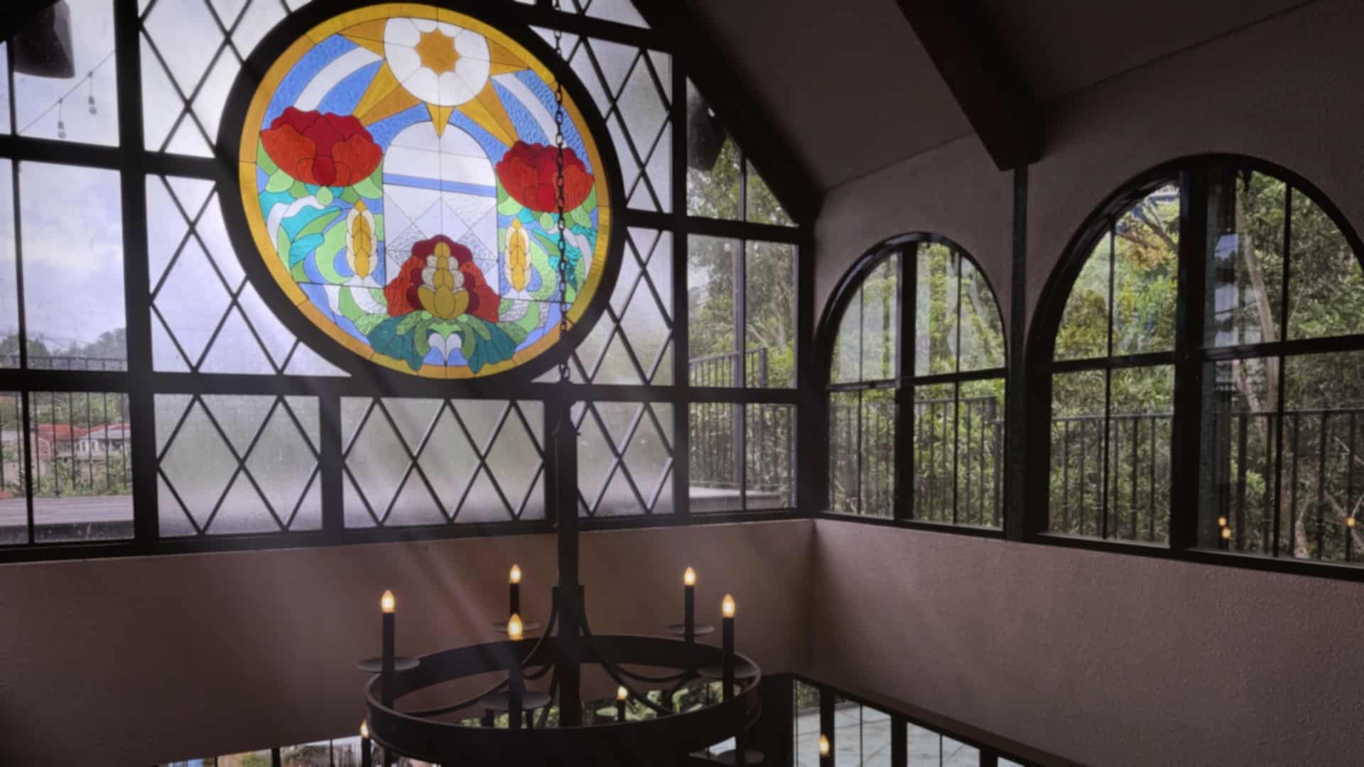 a stained glass window featuring colorful floral designs located above a circular chandelier in a room with large arched windows natural light filters through the glass illuminating the interior digital production A stained glass window featuring colorful floral designs, located above a circular chandelier in a room with large arched windows. Natural light filters through the glass, illuminating the interior.