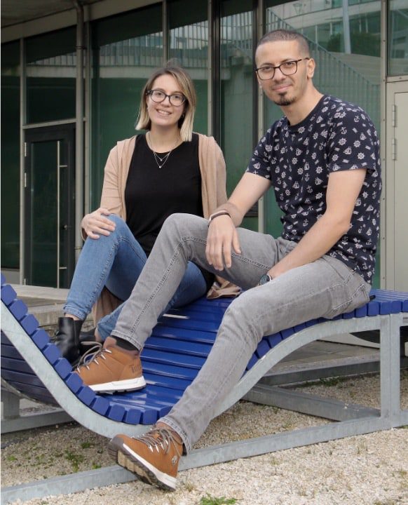 a woman and a man sitting on a blue outdoor bench the woman has short hair and is wearing glasses a black shirt and a cardigan the man has short hair and glasses wearing a patterned shirt and gray pants digital production A woman and a man sitting on a blue outdoor bench. The woman has short hair and is wearing glasses, a black shirt, and a cardigan. The man has short hair and glasses, wearing a patterned shirt and gray pants.