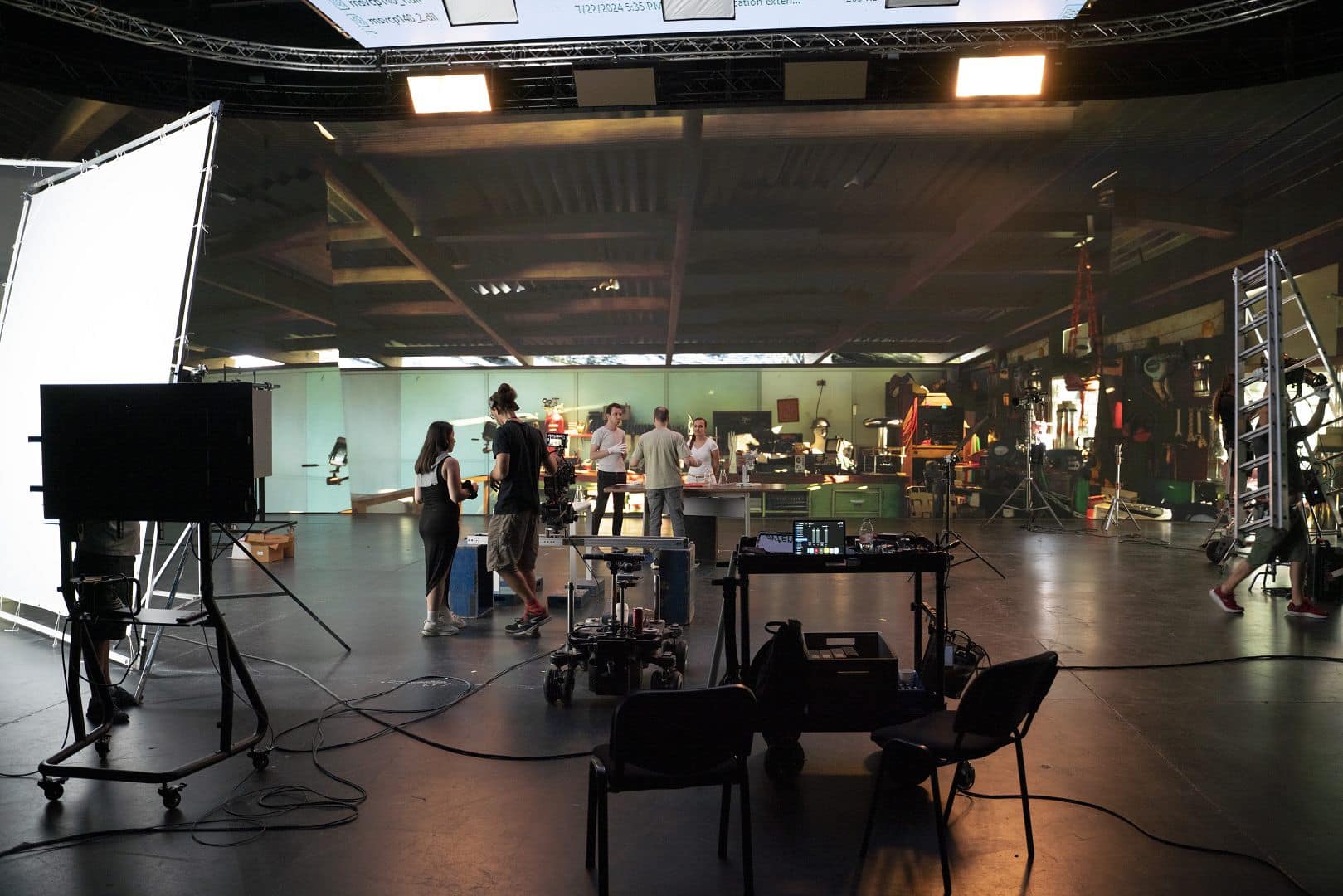 a film set with multiple crew members working in the foreground there are chairs and camera equipment in the background people are gathered near a counter illuminated by studio lights digital production A film set with multiple crew members working. In the foreground, there are chairs and camera equipment. In the background, people are gathered near a counter, illuminated by studio lights.