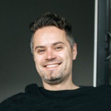 a smiling man with short dark hair wearing a black sweater sits in a modern setting with a neutral background the light casts a warm glow highlighting his cheerful expression digital production A smiling man with short, dark hair, wearing a black sweater, sits in a modern setting with a neutral background. The light casts a warm glow, highlighting his cheerful expression.