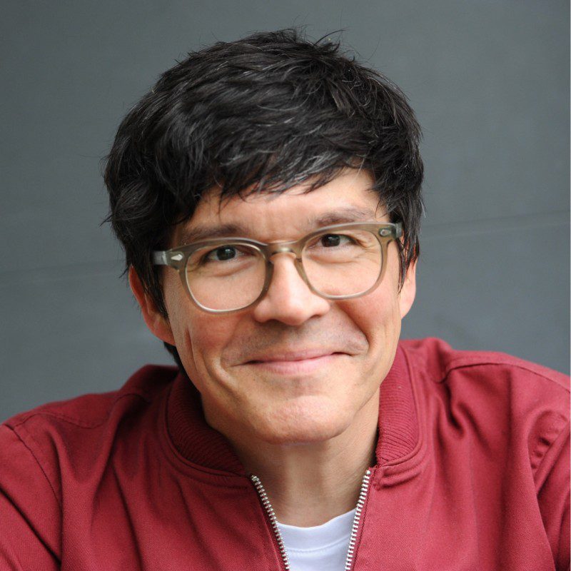 a smiling man with short dark hair and eyeglasses wearing a red jacket over a white shirt he is seated against a gray background conveying a friendly and approachable demeanor digital production A smiling man with short dark hair and eyeglasses, wearing a red jacket over a white shirt. He is seated against a gray background, conveying a friendly and approachable demeanor.