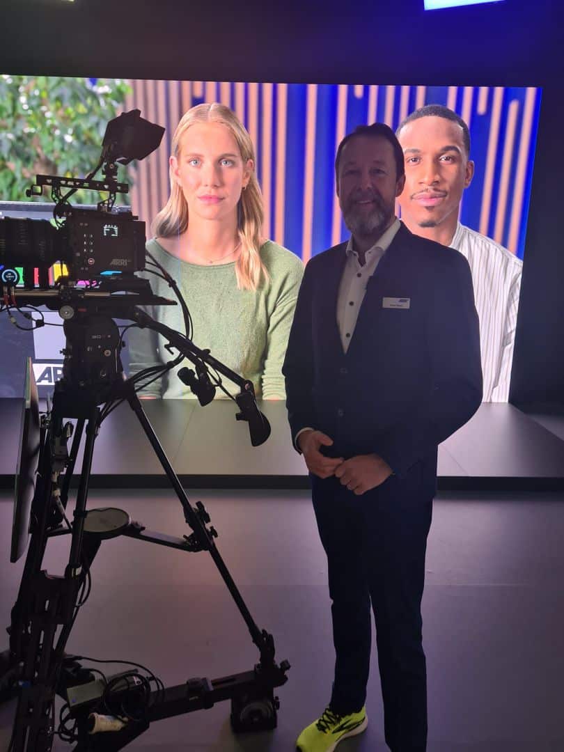 a man in a black suit and bright sneakers stands next to a professional video camera behind him is a large screen displaying two people a woman with long hair in a green sweater and a man in a striped shirt both appearing engaged digital production A man in a suit and sneakers stands beside a video camera on a tripod, with a large screen behind him showing two individuals, one with blonde hair and the other with dark hair, set against a colorful background.