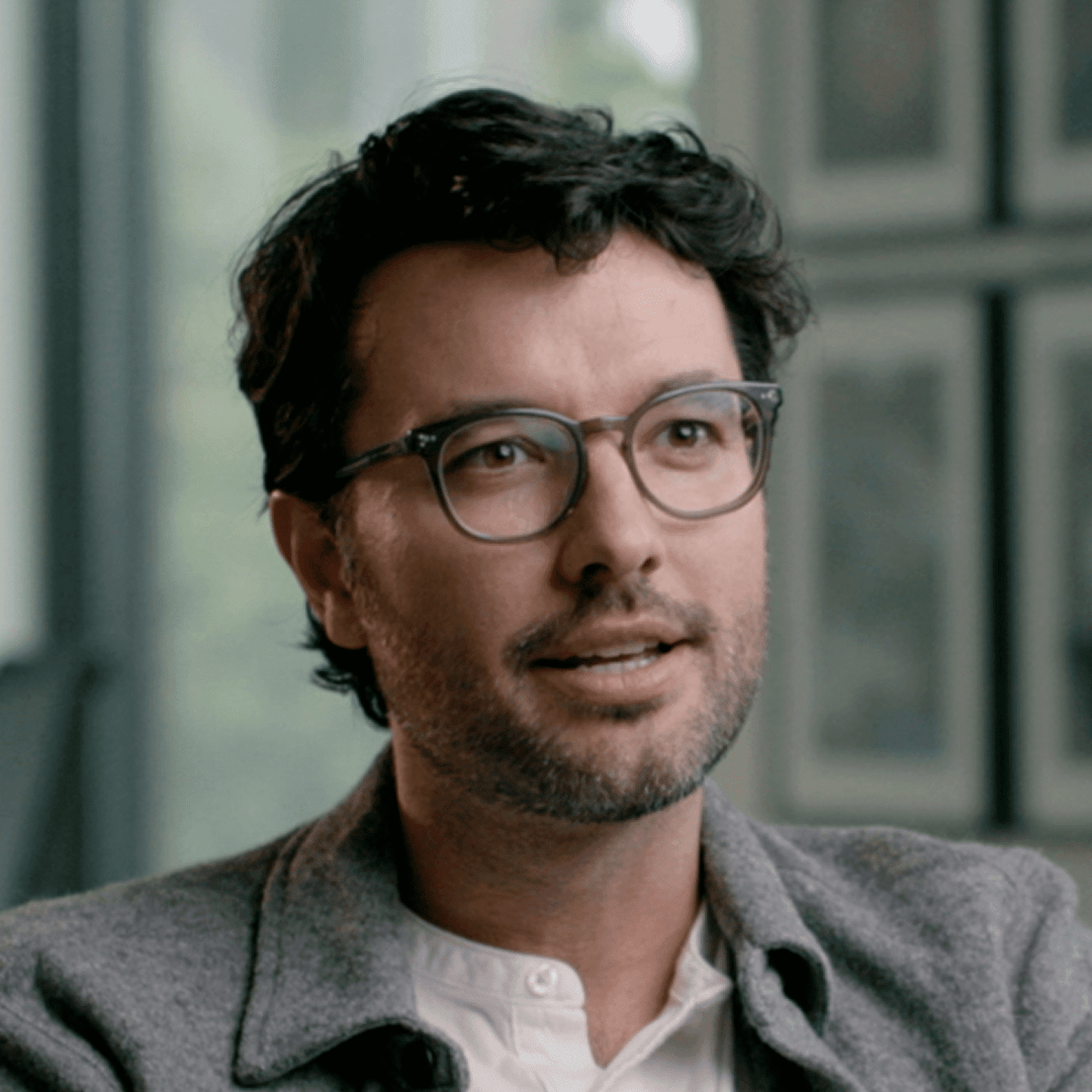 a man with dark hair and glasses speaking thoughtfully in a welllit room with framed pictures in the background he is wearing a lightcolored shirt and a gray jacket appearing engaged in conversation digital production A man with dark hair and glasses speaking thoughtfully in a well-lit room, with framed pictures in the background. He is wearing a light-colored shirt and a gray jacket, appearing engaged in conversation.