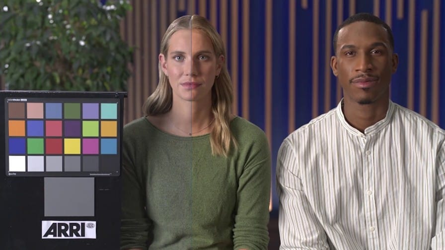 a woman with long hair in a green sweater and a man with short hair in a striped shirt sit beside a color checker board both facing forward a greenery backdrop adds depth to the scene digital production A woman with long hair in a green sweater and a man with short hair in a striped shirt sit beside a color checker board, both facing forward. A greenery backdrop adds depth to the scene.