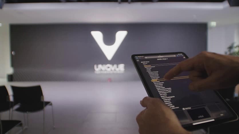 a person using a tablet to navigate a digital interface with a modern promotional stage featuring a large logo reading unovle in the background black chairs are arranged in front of the stage digital production A person using a tablet to navigate a digital interface, with a modern promotional stage featuring a large logo reading "UNOVLE" in the background. Black chairs are arranged in front of the stage.