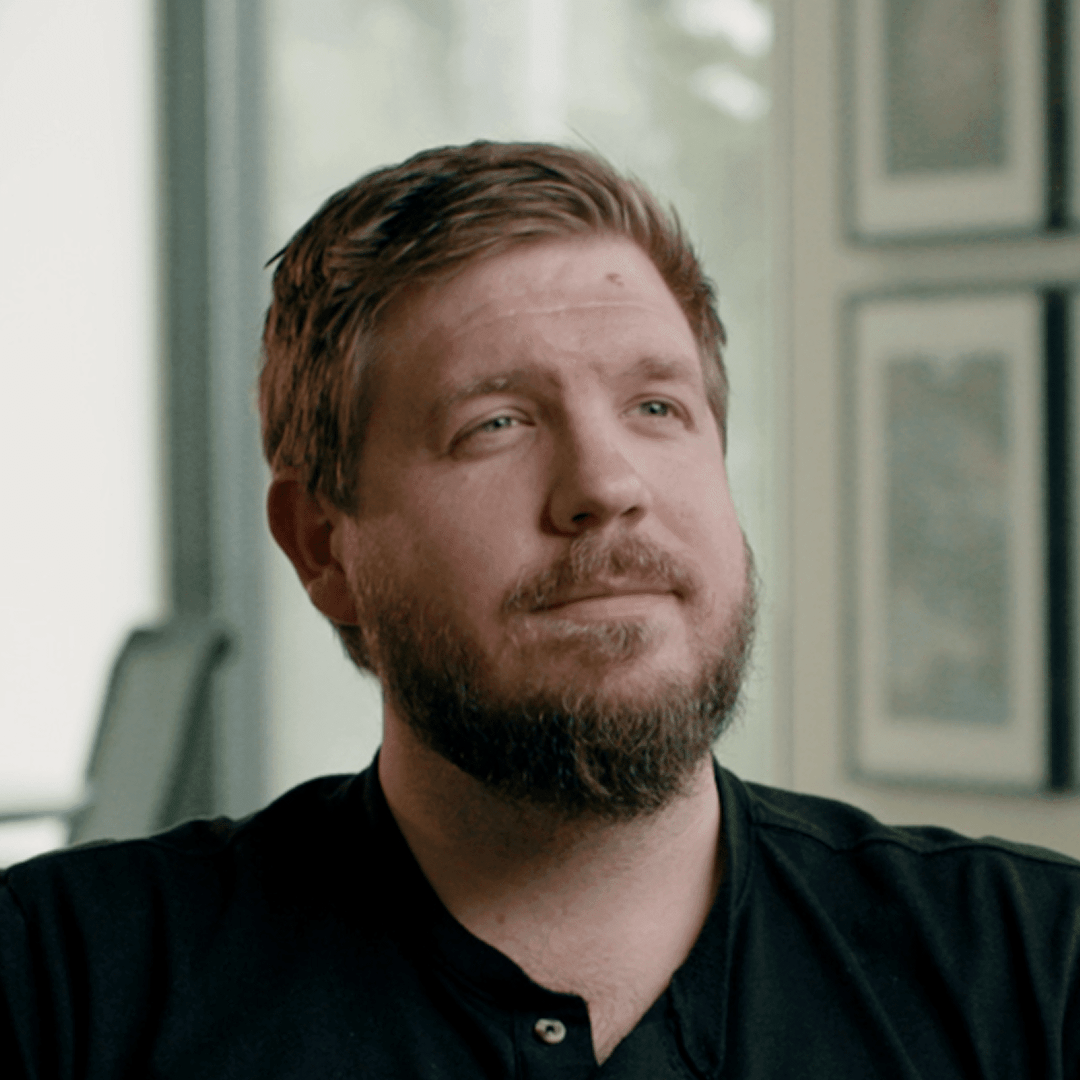 a man with a beard and short hair sits in a welllit room looking thoughtfully towards the right he is wearing a black shirt and there are framed pictures on the walls in the background digital production A man with a beard and short hair sits in a well-lit room, looking thoughtfully towards the right. He is wearing a black shirt, and there are framed pictures on the walls in the background.