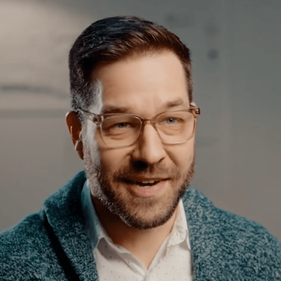 a smiling man with a beard and glasses wearing a blue cardigan over a white shirt sits in a welllit indoor setting appearing engaged in conversation digital production A smiling man with a beard and glasses, wearing a blue cardigan over a white shirt, sits in a well-lit indoor setting, appearing engaged in conversation.