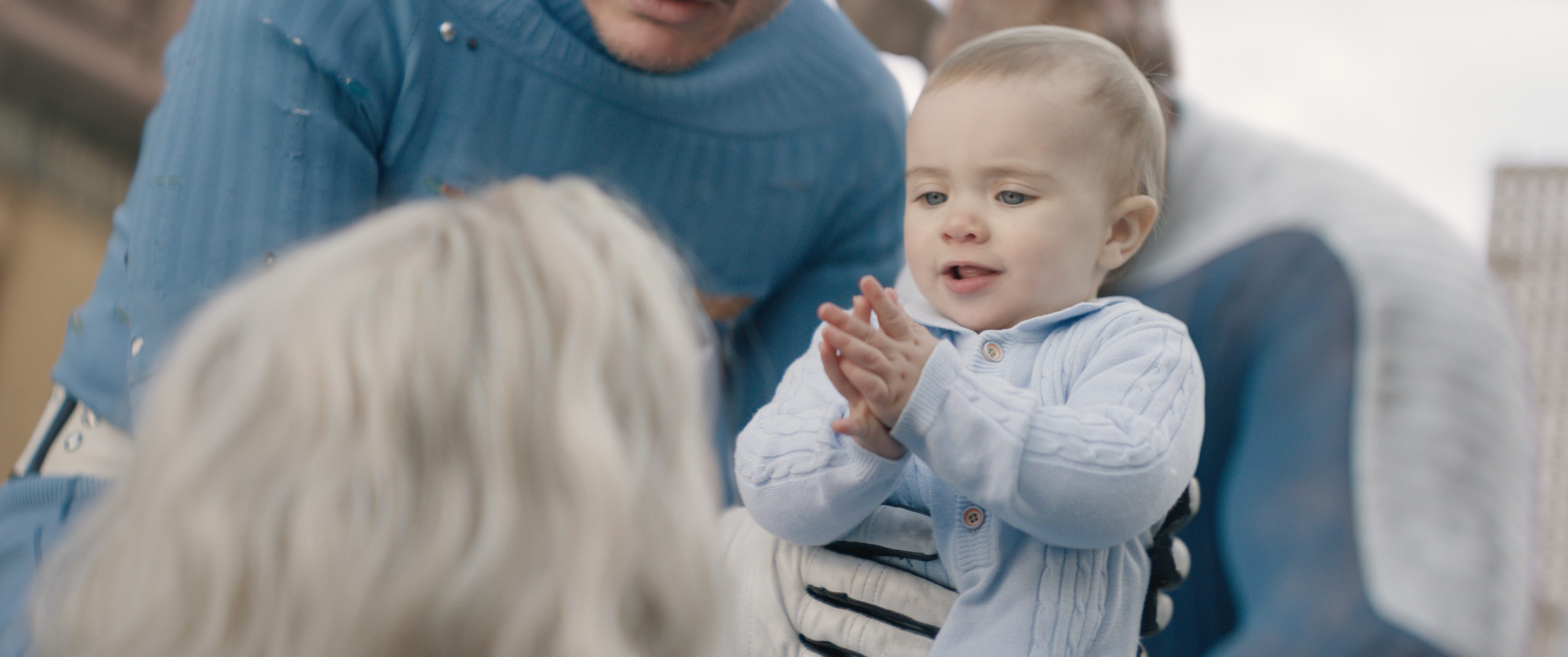 a baby in a light blue sweater claps happily while looking at an adult in a blue costume the background shows a blurred figure suggesting a cheerful outdoor scene digital production A baby in a light blue sweater claps happily while looking at an adult in a blue costume. The background shows a blurred figure, suggesting a cheerful outdoor scene.