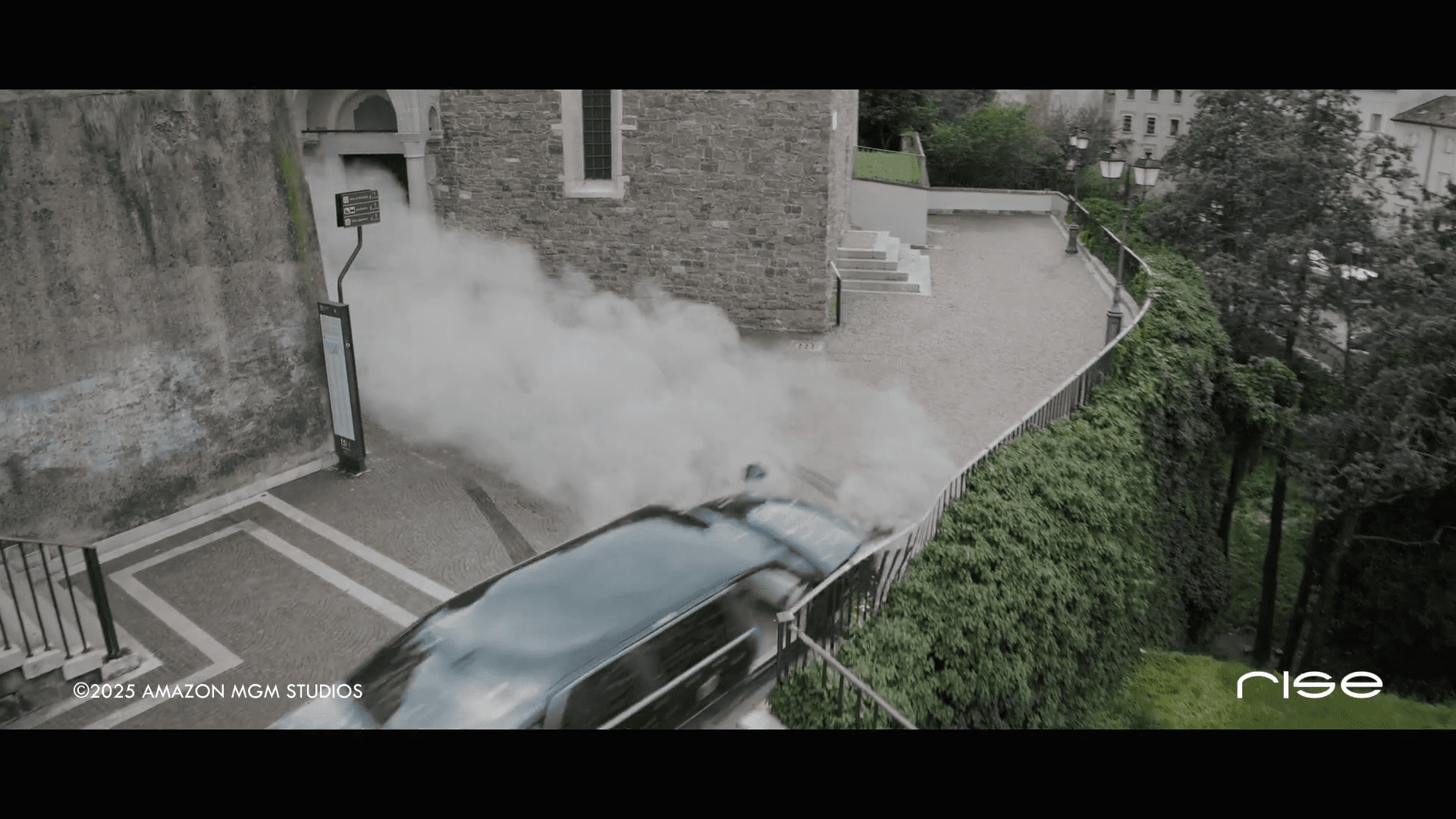 a black car drives off a winding road near a stone building releasing a cloud of smoke as it accelerates around a bend with greenery and stairs visible on the side digital production A black car drives off a winding road near a stone building, releasing a cloud of smoke as it accelerates around a bend, with greenery and stairs visible on the side.