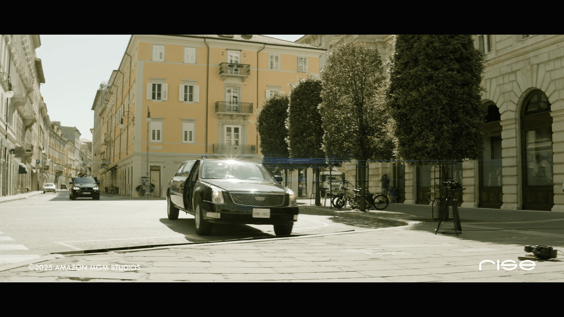 a black sedan drives down a sunny deserted street lined with trees and classic buildings a bicycle and a camera equipment setup are visible on the sidewalk hinting at a film shoot digital production A black sedan drives down a sunny, deserted street lined with trees and classic buildings. A bicycle and a camera equipment setup are visible on the sidewalk, hinting at a film shoot.
