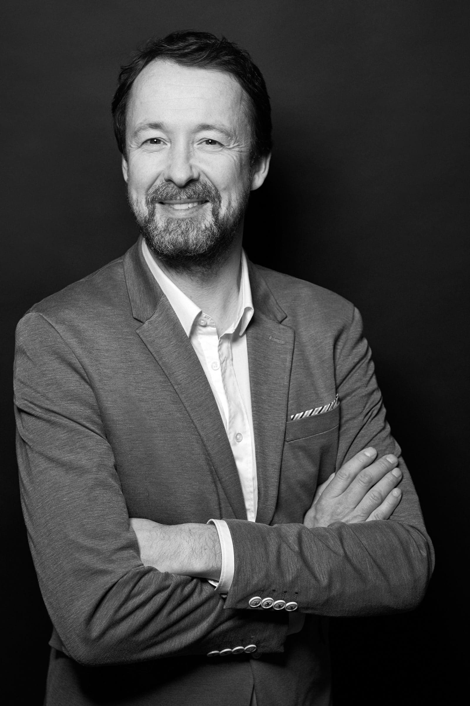 a man with a beard wearing a gray blazer and white shirt smiles confidently with arms crossed against a solid black background the photo is in black and white emphasizing the subjects friendly demeanor and professional appearance digital production A man with a beard smiles confidently while crossing his arms, wearing a light gray blazer over a white shirt. The background is dark, emphasizing his expression and attire.