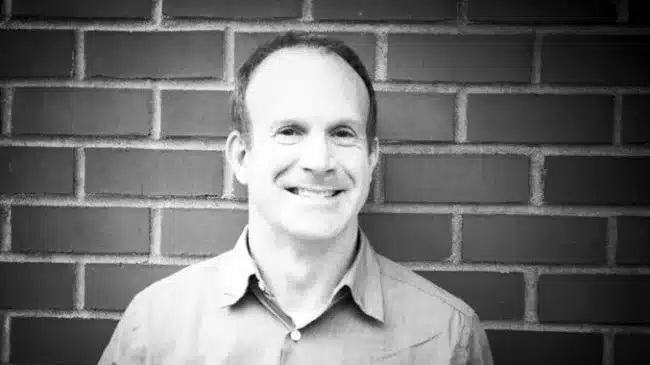 a man smiling in front of a brick wall wearing a lightcolored shirt the photo has a black and white filter giving it a classic feel digital production A man smiling in front of a brick wall, wearing a light-colored shirt. The photo has a black and white filter, giving it a classic feel.