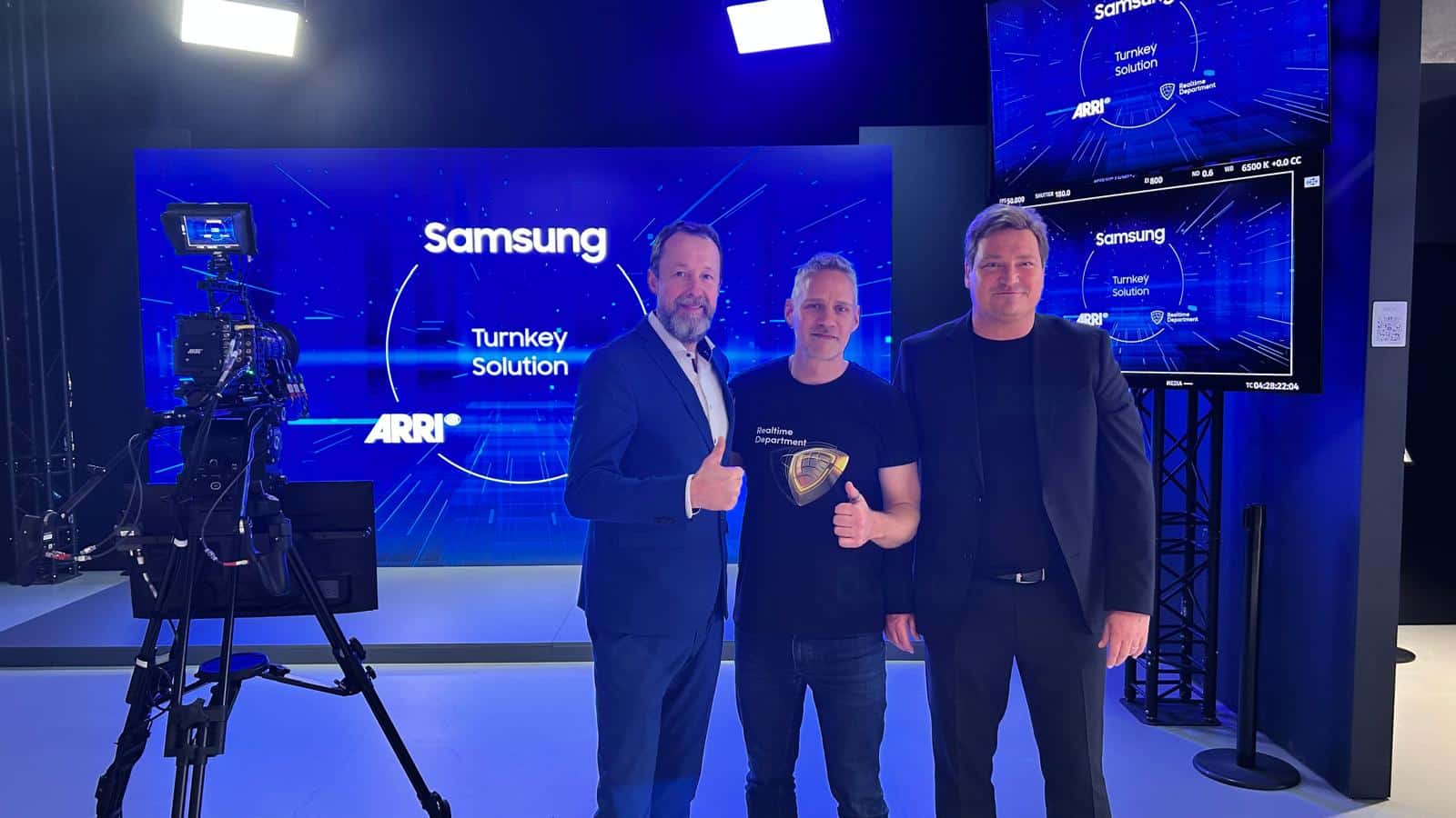 three men posing together on stage at a technology event they are smiling and giving thumbs up behind them there is a screen displaying the samsung logo and the words turnkey solution with blue graphic elements digital production Three professionals standing in a studio environment, posing with thumbs up. A camera is visible in the foreground, with a blue background displaying the Samsung logo and text related to a "Turnkey Solution."