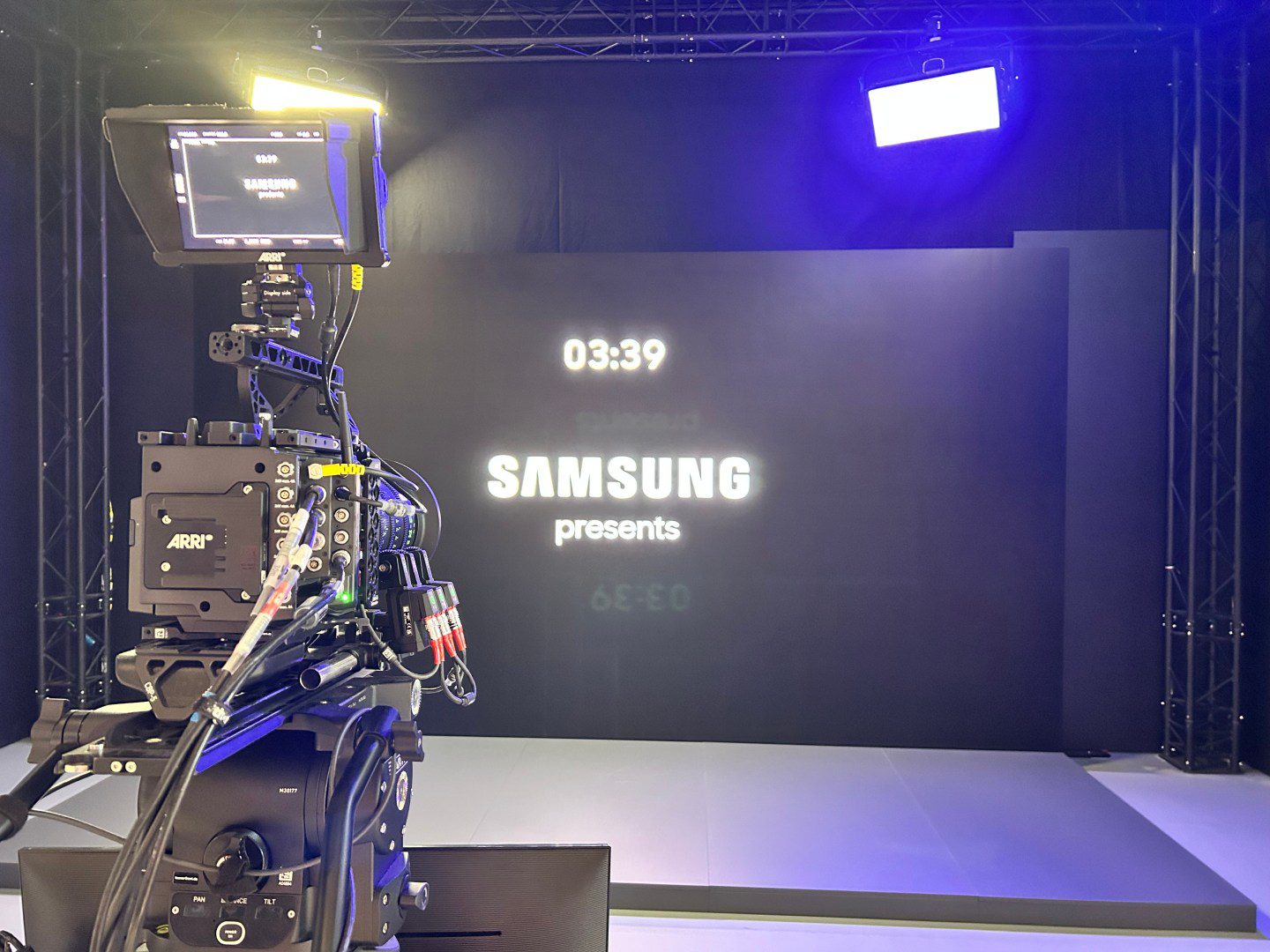 a professional camera set up in a studio focused on a digital screen displaying a countdown timer 0339 and the text samsung presents in bold white letters against a dark background digital production A professional camera setup in a studio, focused on a backdrop displaying 'Samsung presents' with a countdown timer showing 03:39. Bright stage lights illuminate the scene.