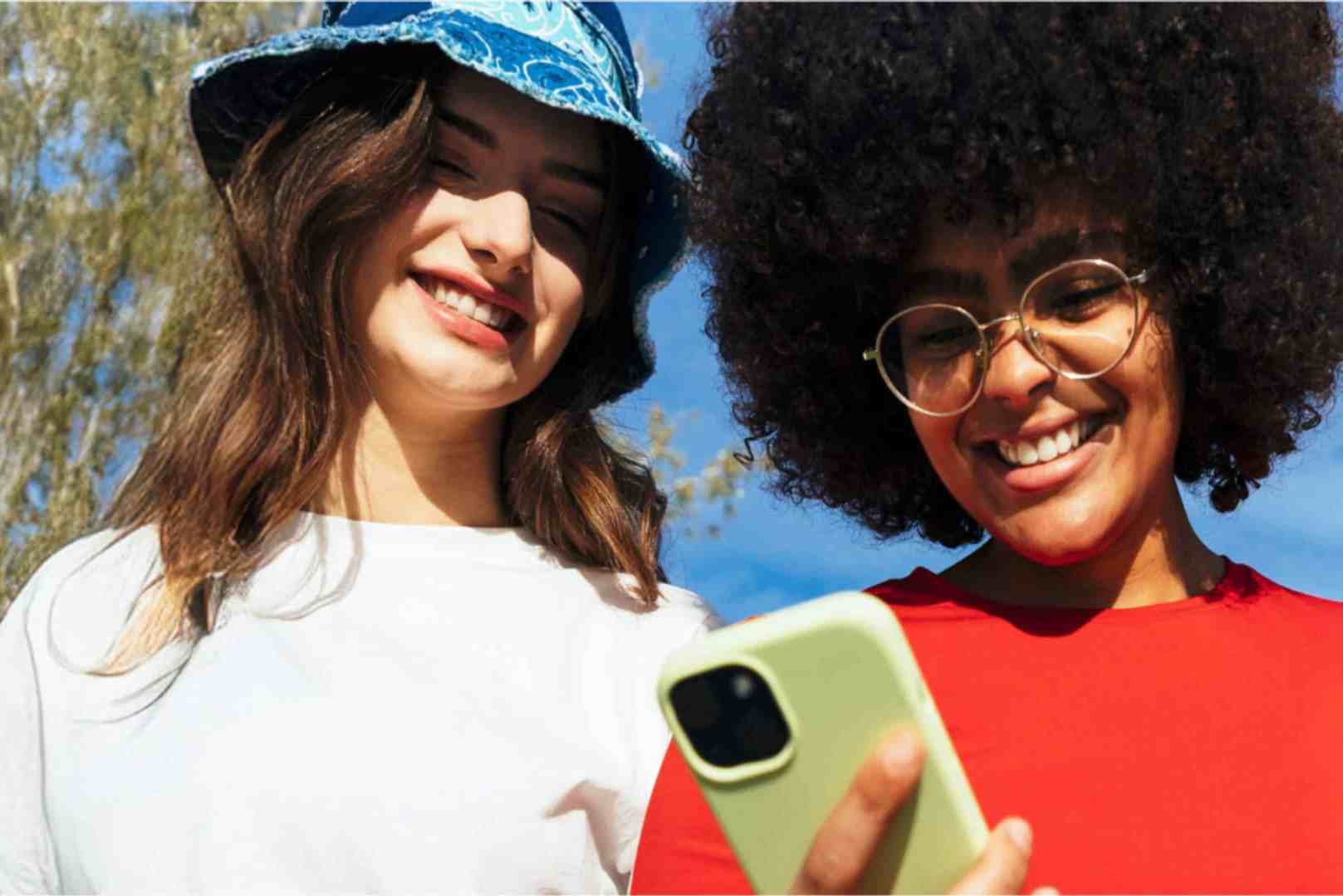 two young women outdoors one in a white shirt and blue bucket hat the other in a red shirt with curly hair are smiling while looking at a smartphone together bright blue sky and trees are in the background digital production Two young women outdoors, one in a white shirt and blue bucket hat, the other in a red shirt with curly hair, are smiling while looking at a smartphone together. Bright blue sky and trees are in the background.