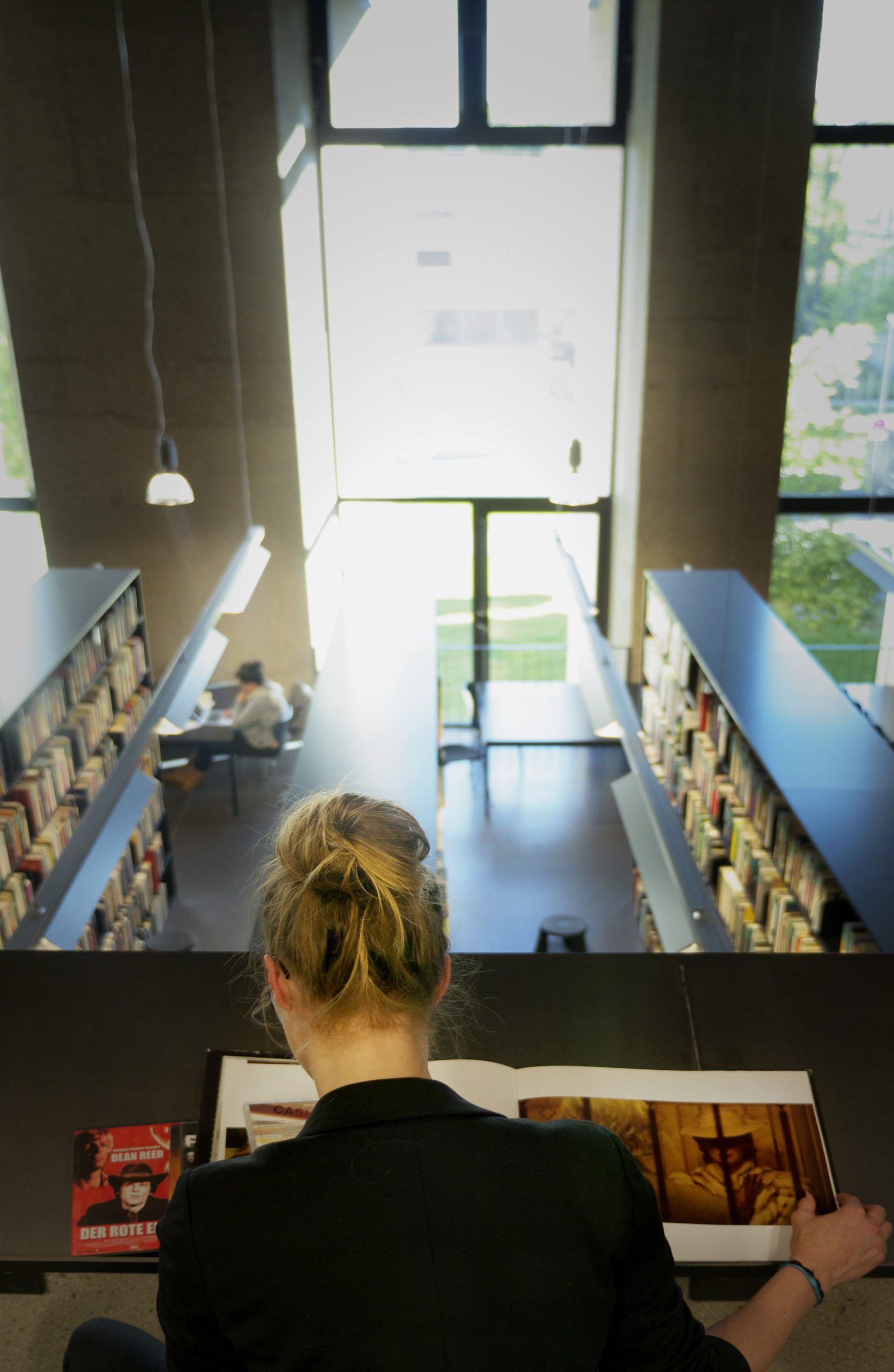 a woman with her back to the camera sits at a desk studying a large book in a modern library natural light streams through large windows illuminating the shelves filled with books and a person seated in the background digital production A woman with her back to the camera sits at a desk, studying a large book, in a modern library. Natural light streams through large windows, illuminating the shelves filled with books and a person seated in the background.