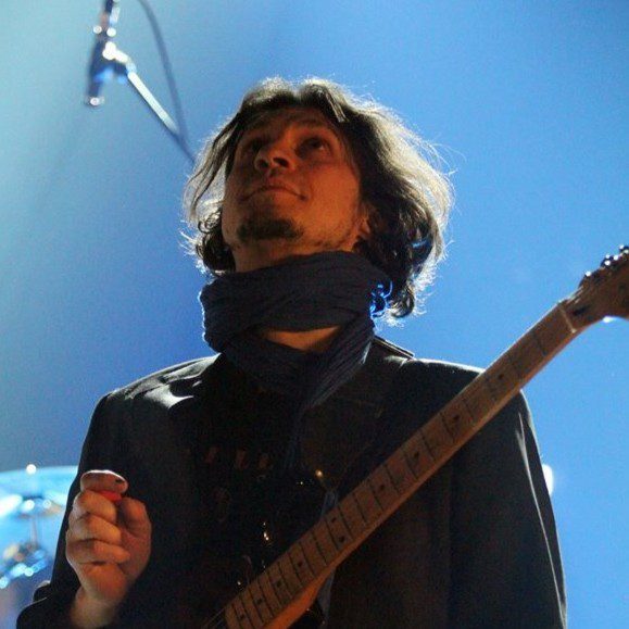 a musician playing guitar on stage gazing upwards under soft blue lighting he has tousled hair and wears a scarf and a dark jacket creating an artistic ambiance digital production A musician playing guitar on stage, gazing upwards under soft blue lighting. He has tousled hair and wears a scarf and a dark jacket, creating an artistic ambiance.