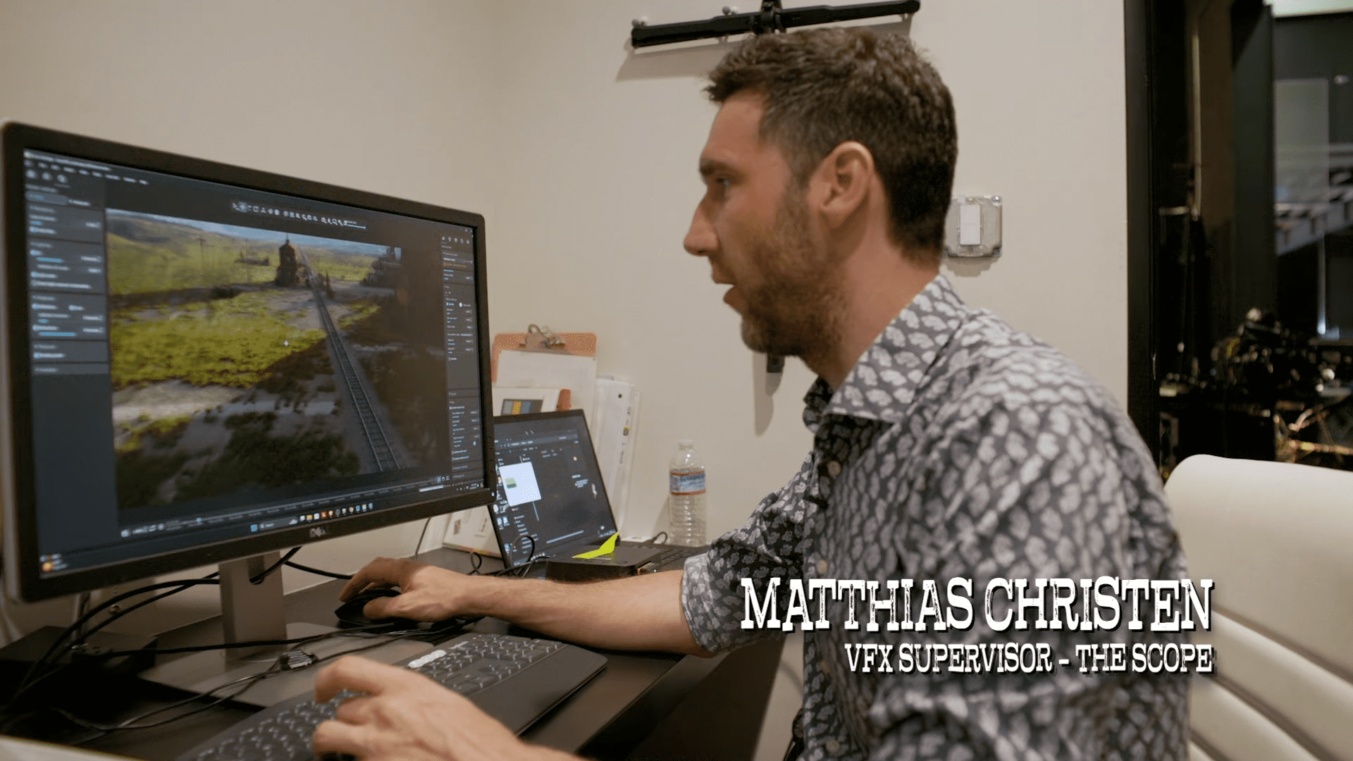 a man with short hair and a patterned shirt focused on his computer screen in an office setting the screen displays a landscape image while he navigates digital editing software surrounded by office equipment digital production A man with short hair and a patterned shirt focused on his computer screen in an office setting. The screen displays a landscape image, while he navigates digital editing software, surrounded by office equipment.