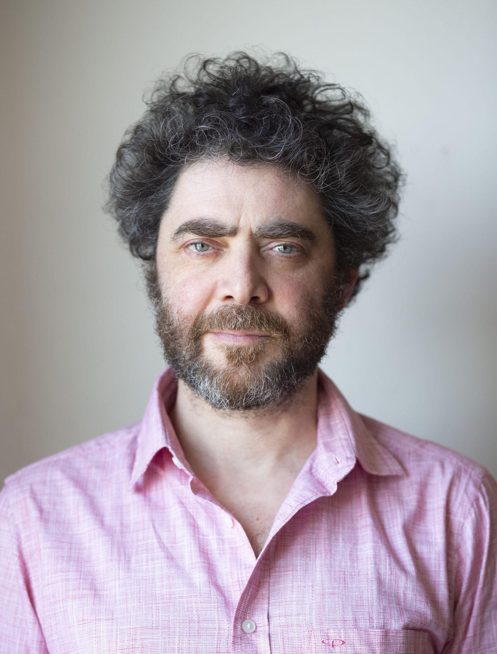 © ania gruca digital production A man with curly dark hair and a beard, wearing a light pink shirt, stands against a simple background. He looks directly at the camera with a serious expression, the soft lighting highlighting his facial features.