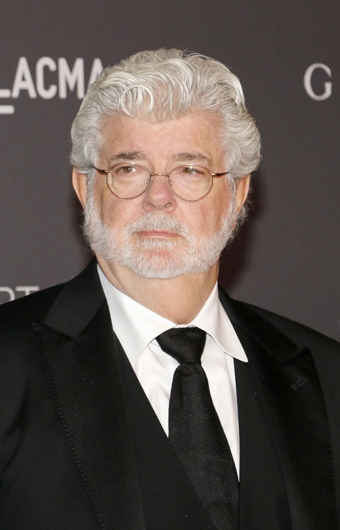 a man with wavy gray hair and a beard wearing glasses dressed in a black tuxedo and white shirt poses for the camera at an elegant event with a dark background digital production A man with wavy gray hair and a beard wearing glasses, dressed in a black tuxedo and white shirt, poses for the camera at an elegant event with a dark background.