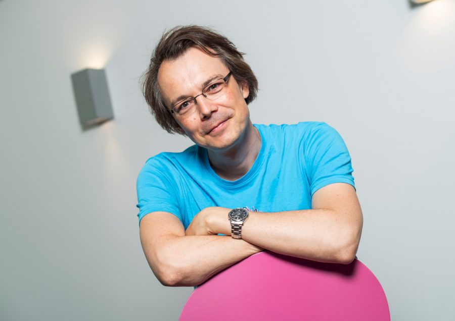 a man with light brown hair and glasses smiles while leaning on a large pink object he wears a bright blue shirt and has a relaxed posture in a modern softly lit space digital production A man with light brown hair and glasses smiles while leaning on a large pink object. He wears a bright blue shirt and has a relaxed posture in a modern, softly lit space.