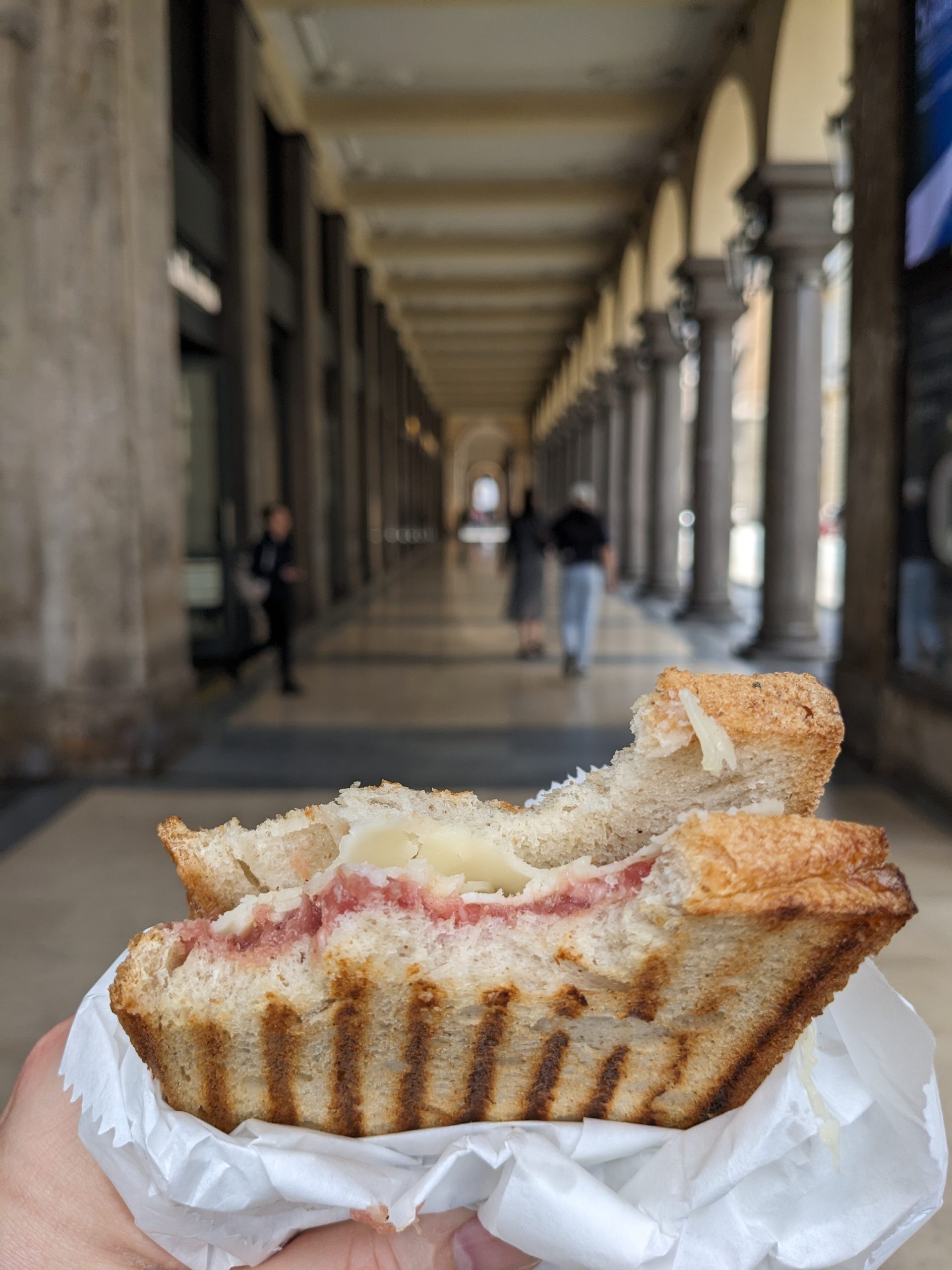 a hand holding a halfeaten sandwich with a toasted exterior filled with layers of ham and cheese in a long arcade with arches in the background people walk in the distance along the corridor digital production A hand holding a half-eaten sandwich with a toasted exterior, filled with layers of ham and cheese, in a long arcade with arches in the background. People walk in the distance along the corridor.
