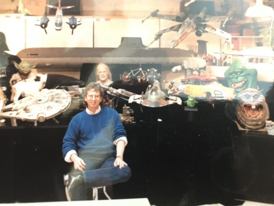 a smiling man sitting on a chair in a workshop surrounded by various collectible figures including spaceships and characters from science fiction movies bright overhead lights illuminate the eclectic assortment of memorabilia digital production A smiling man sitting on a chair in a workshop surrounded by various collectible figures including spaceships and characters from science fiction movies. Bright overhead lights illuminate the eclectic assortment of memorabilia.