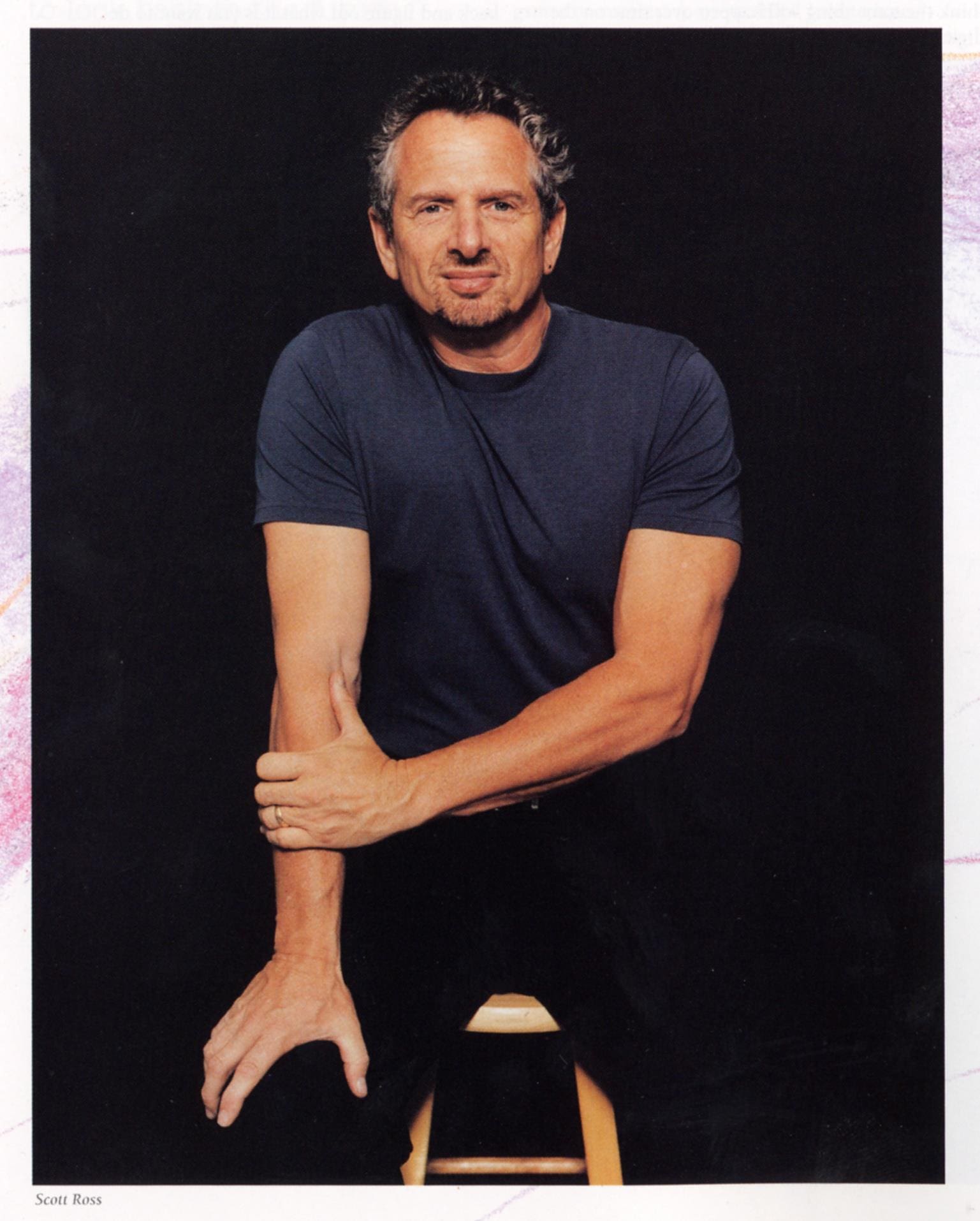 a man with curly gray hair and a beard sits on a stool wearing a dark blue tshirt and black pants he rests one arm on his knee while placing his other hand on his thigh set against a black background digital production A man with curly gray hair and a beard sits on a stool, wearing a dark blue t-shirt and black pants. He rests one arm on his knee while placing his other hand on his thigh, set against a black background.