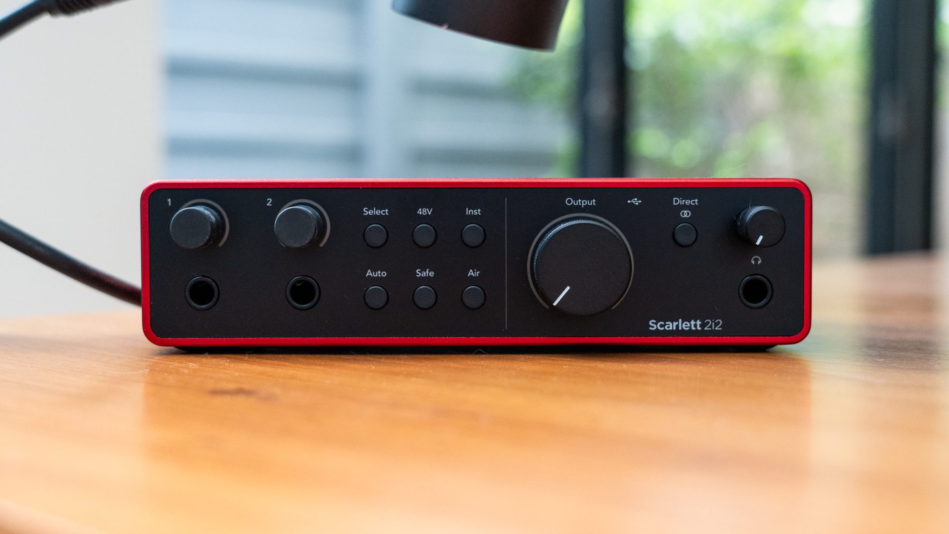 a closeup view of a scarlett 2i2 audio interface featuring various knobs and buttons resting on a wooden table the interface is predominantly black with red accents with clear labels and controls for audio output and input settings digital production A close-up view of a Scarlett 2i2 audio interface, featuring various knobs and buttons, resting on a wooden table. The interface is predominantly black with red accents, with clear labels and controls for audio output and input settings.
