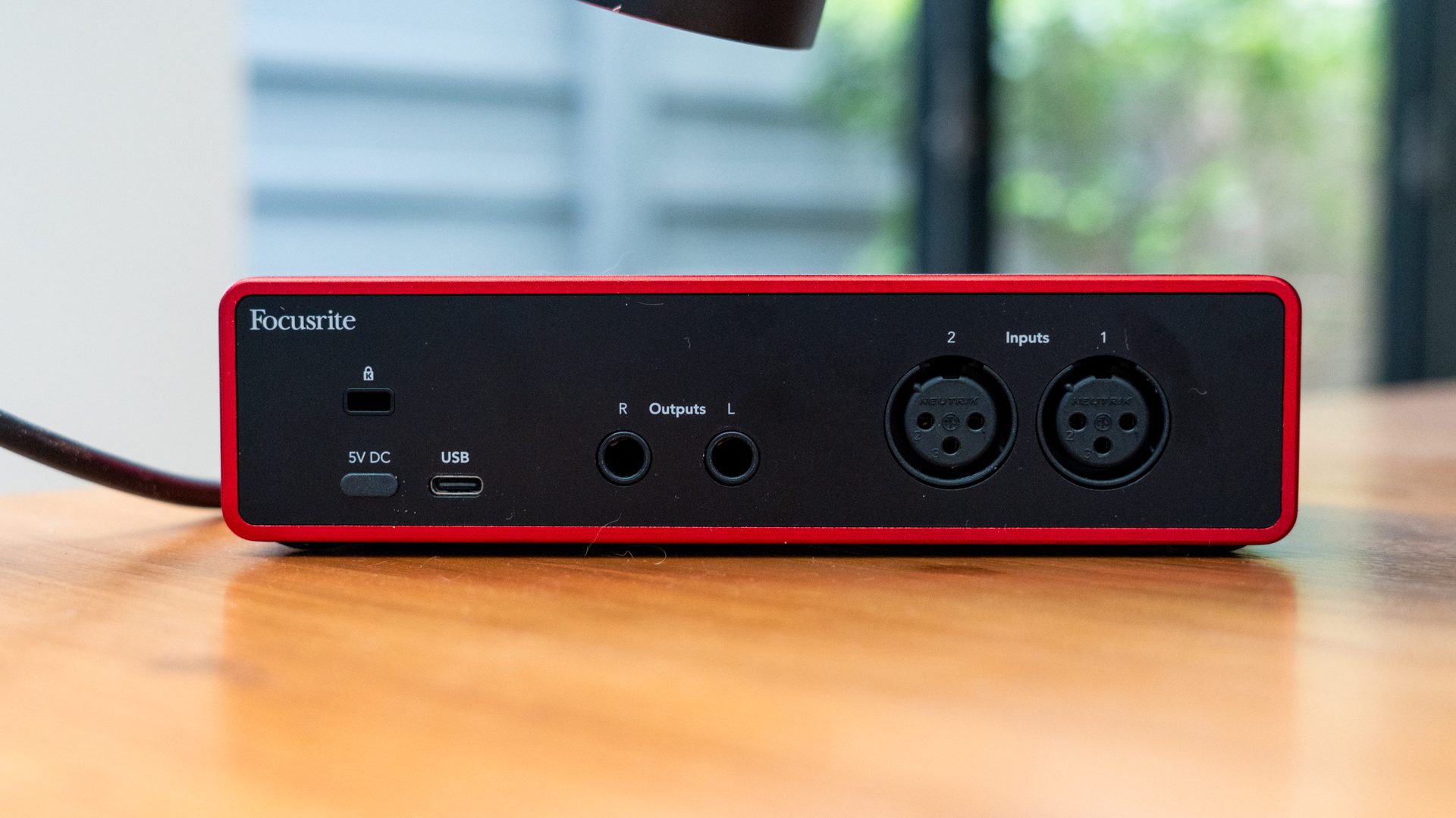 a closeup view of a focusrite audio interface featuring a red and black design the front panel includes input and output ports a usb connection and indicator lights set on a wooden table with a blurred background digital production A close-up view of a Focusrite audio interface featuring a red and black design. The front panel includes input and output ports, a USB connection, and indicator lights, set on a wooden table with a blurred background.