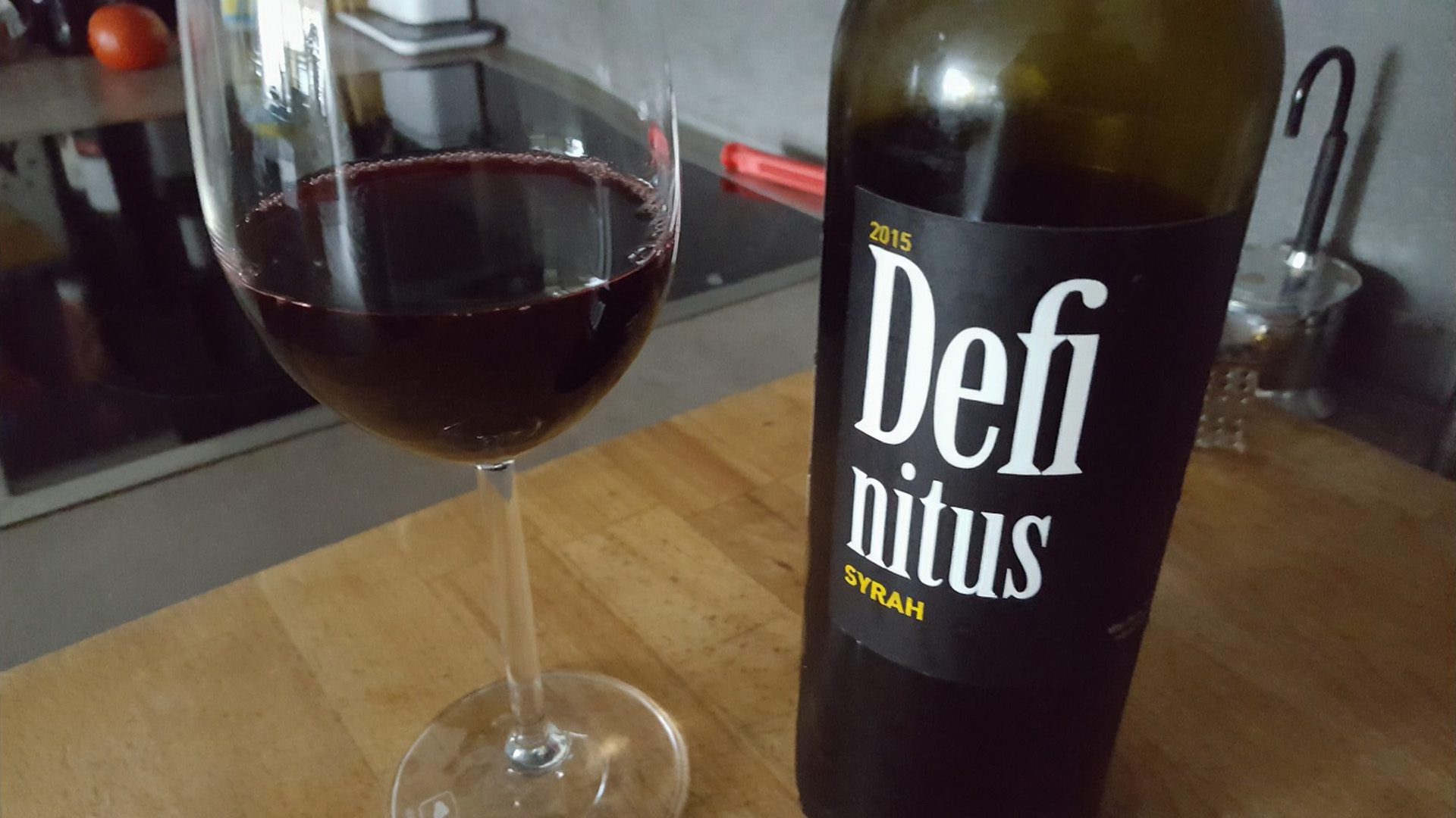 a filled wine glass next to a bottle of definitus syrah from 2015 placed on a wooden surface the glass displays deep red wine reflecting light while the bottle features a bold black label digital production A filled wine glass next to a bottle of Definitus Syrah from 2015, placed on a wooden surface. The glass displays deep red wine reflecting light, while the bottle features a bold black label.