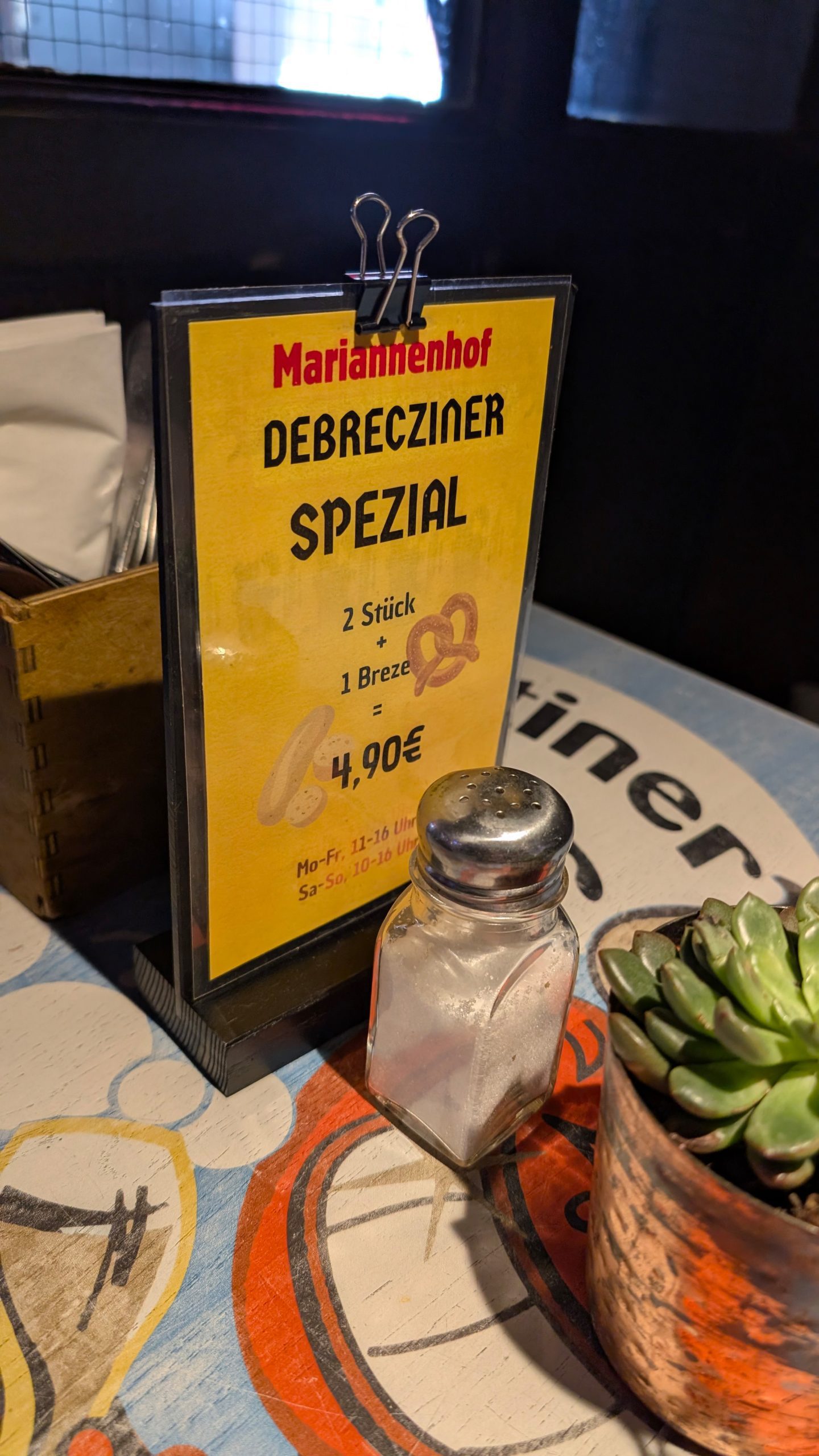 a menu stand featuring a yellow and red sign that reads debrecziner spezial with items and prices including 2 stück and 1 breze for 490€ on a colorful table with a salt shaker and a small succulent digital production A menu stand featuring a yellow and red sign that reads 'Debrecziner Spezial' with items and prices, including '2 Stück' and '1 Breze' for 4,90€ on a colorful table with a salt shaker and a small succulent.