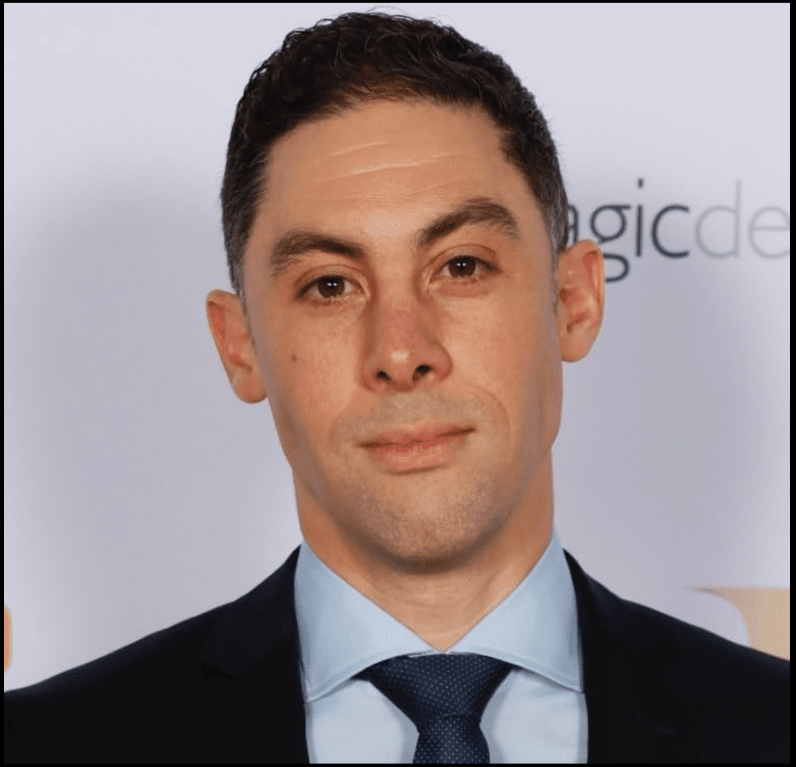 a closeup portrait of a man in a formal suit featuring a light blue dress shirt and a dark tie he has short dark hair and a neutral expression with a blurred background displaying a logo digital production A close-up portrait of a man in a formal suit, featuring a light blue dress shirt and a dark tie. He has short dark hair and a neutral expression, with a blurred background displaying a logo.